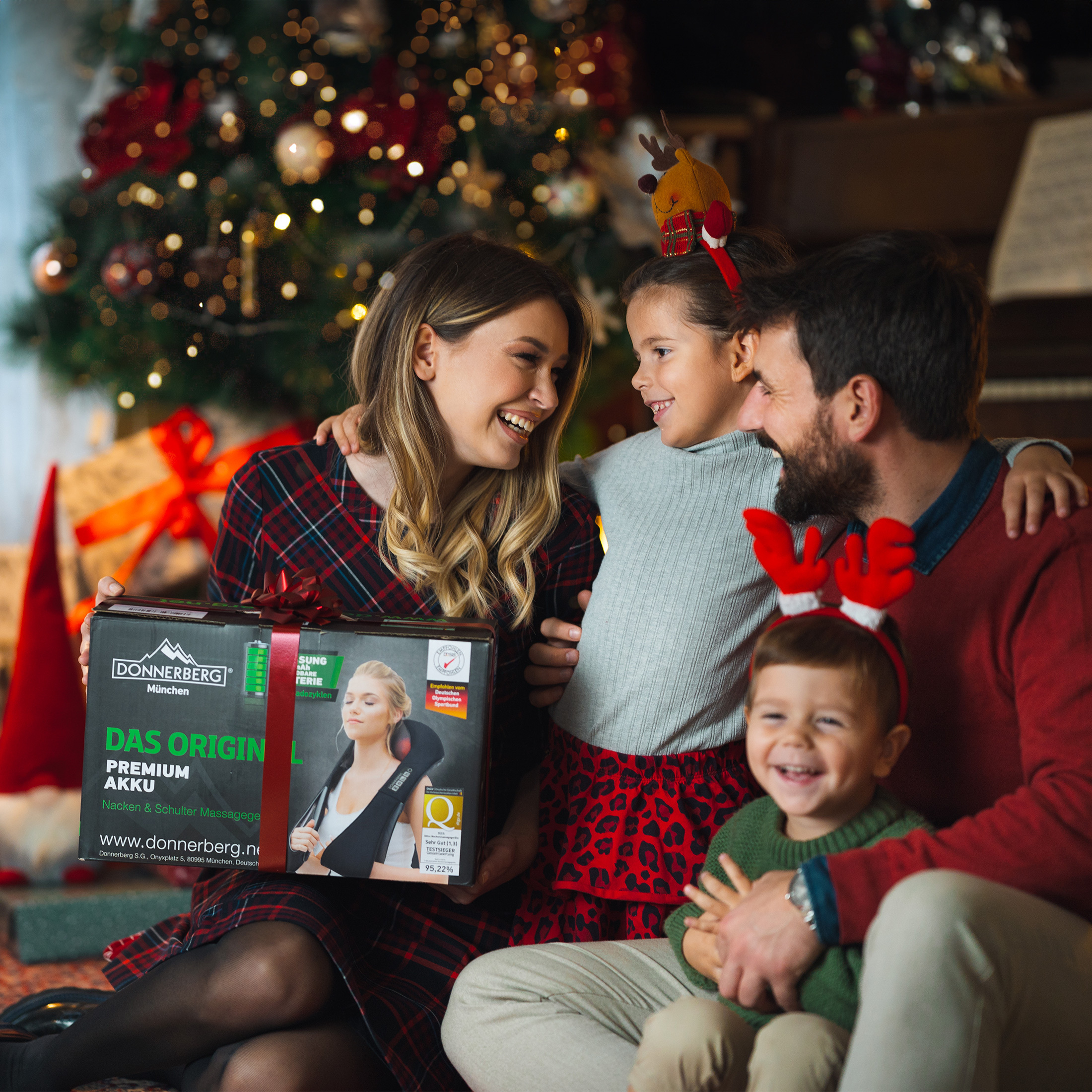 Eine Familie in einem Raum mit Weihnachtsbaum. Sie halten eine Geschenkbox mit Text darauf.