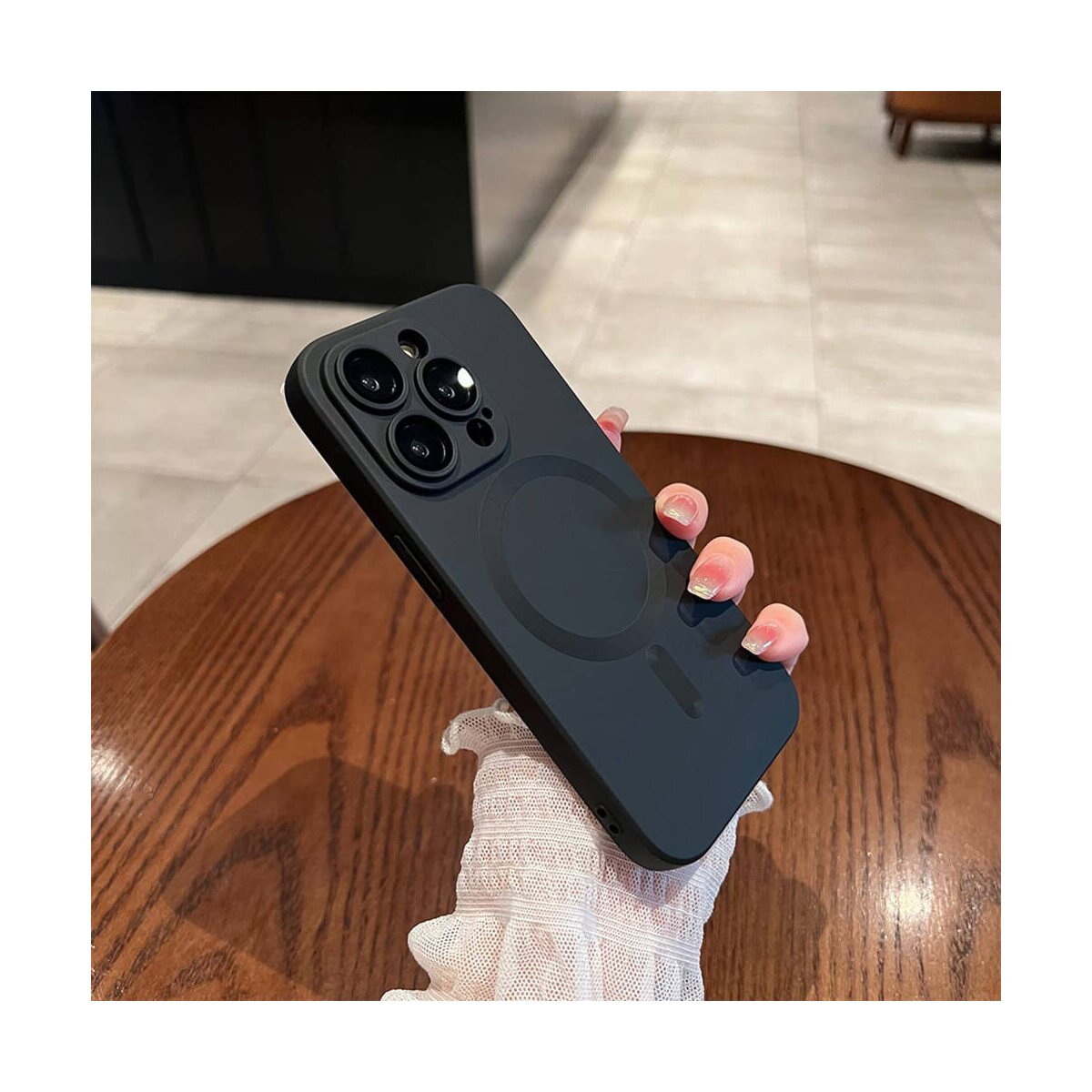 Una mano che tiene una custodia nera per telefono. La custodia ha un cerchio MagSafe. Tavolo di legno.
