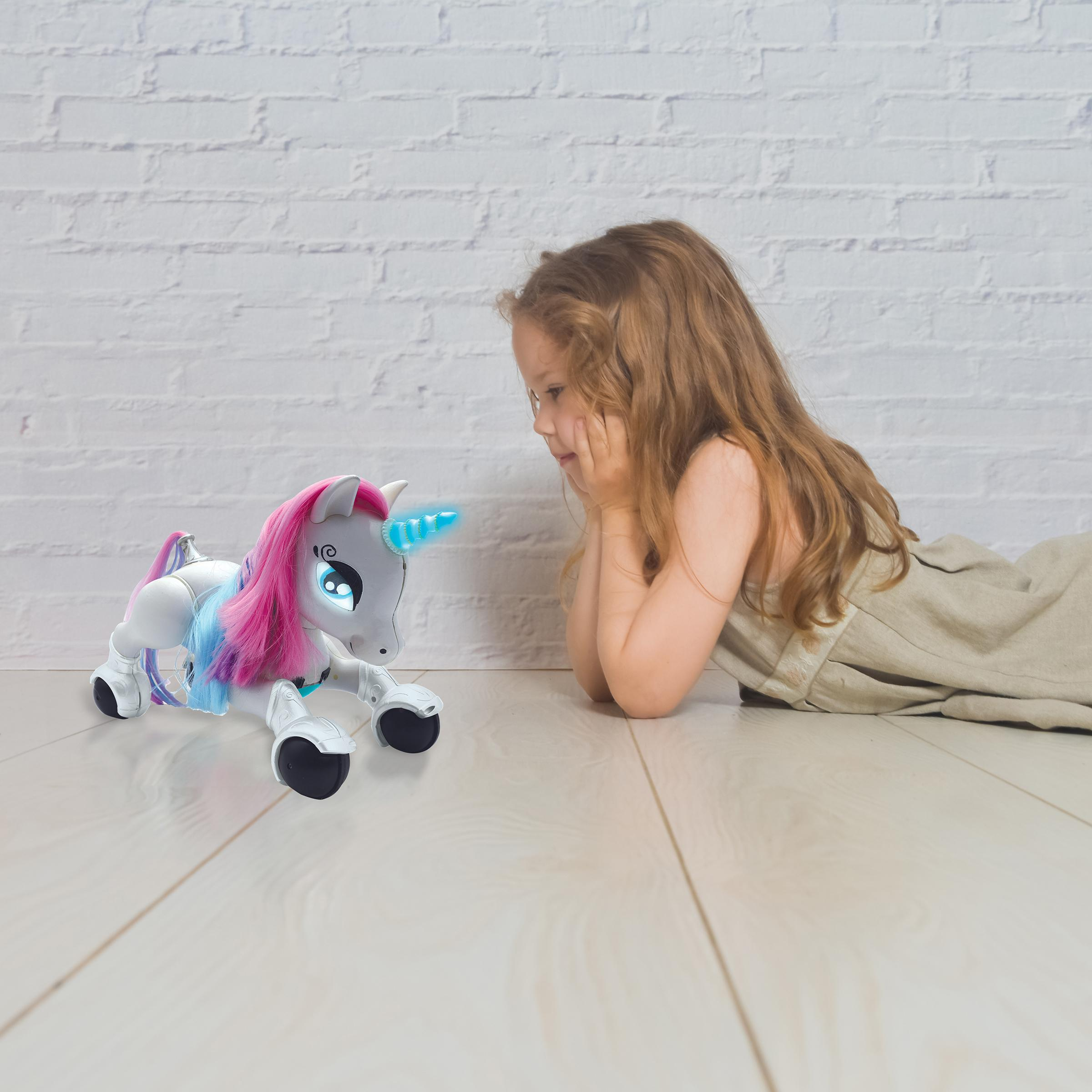 Bambina guarda unicorno giocattolo su pavimento legno, muro mattoni bianchi.