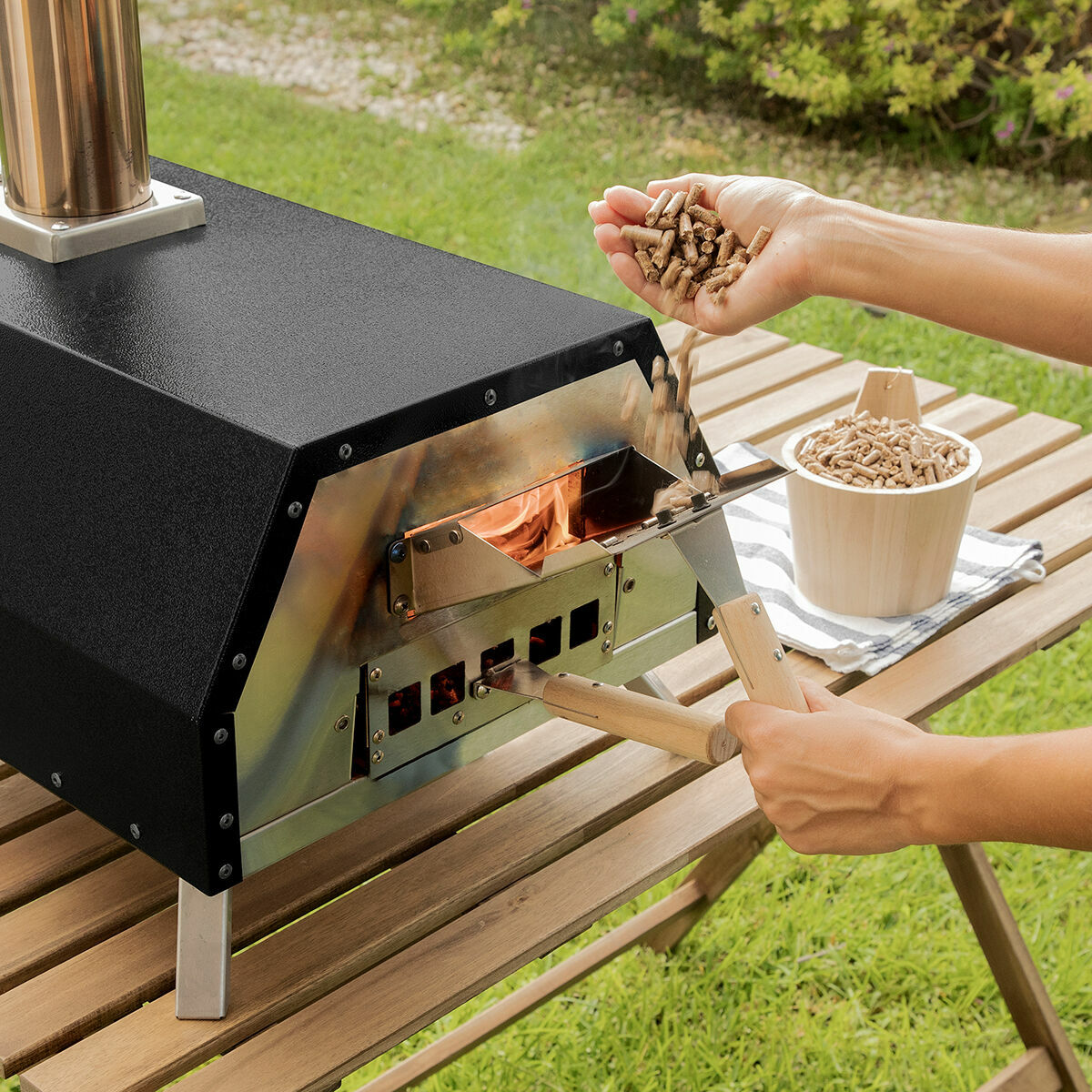 Forno per pizza nero all'aperto con una persona che aggiunge pellet di legno. Erba verde.