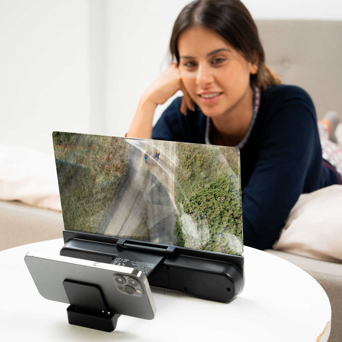 Mujer mirando una pantalla de teléfono ampliada por un dispositivo negro en una mesa blanca.
