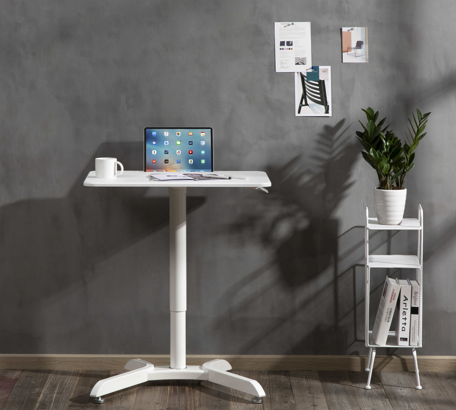 Een witte statafel met een tablet, een mok, boeken en een plant tegen een grijze muur.