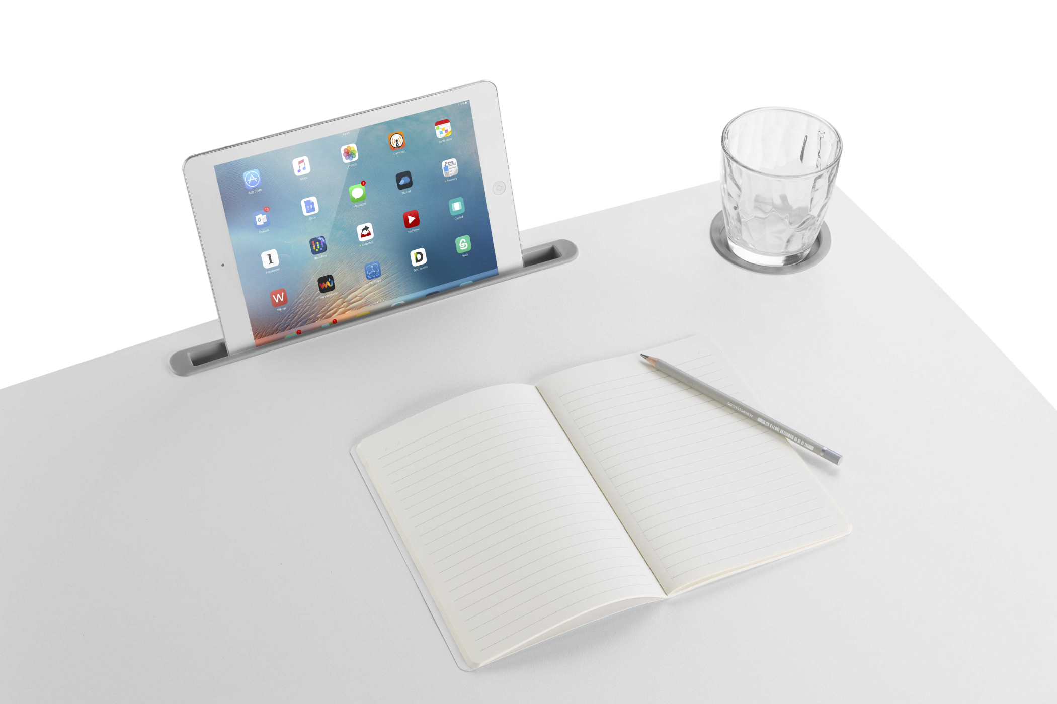 Tablet in een bureausleuf, notitieboek, potlood en glas op een wit bureau.