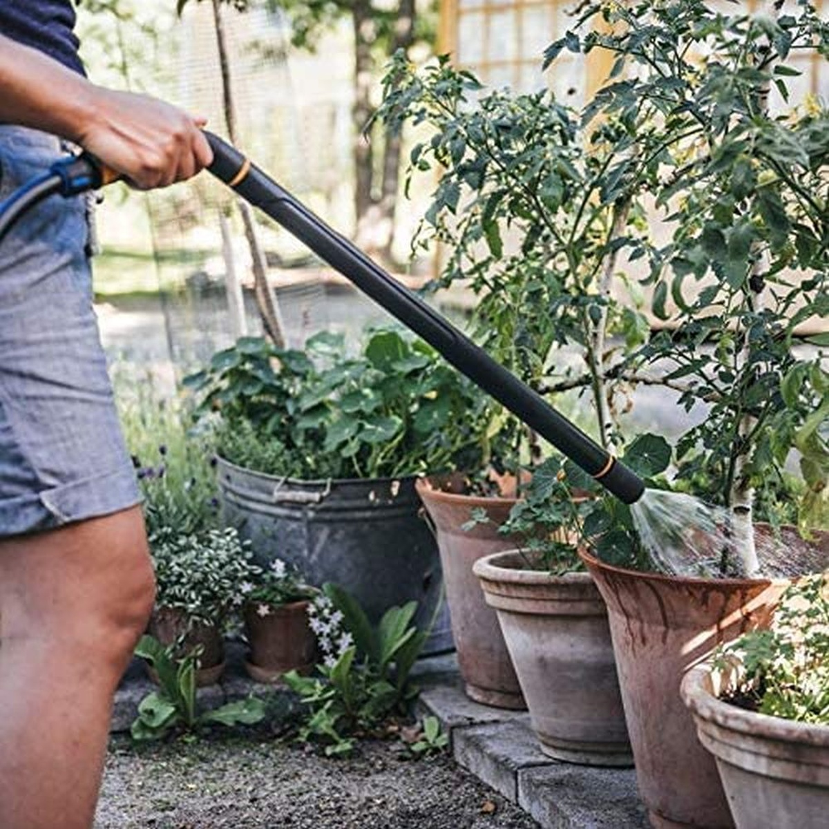 Eine Person gießt Pflanzen mit einem Schlauch. Topfpflanzen befinden sich in einem Garten.