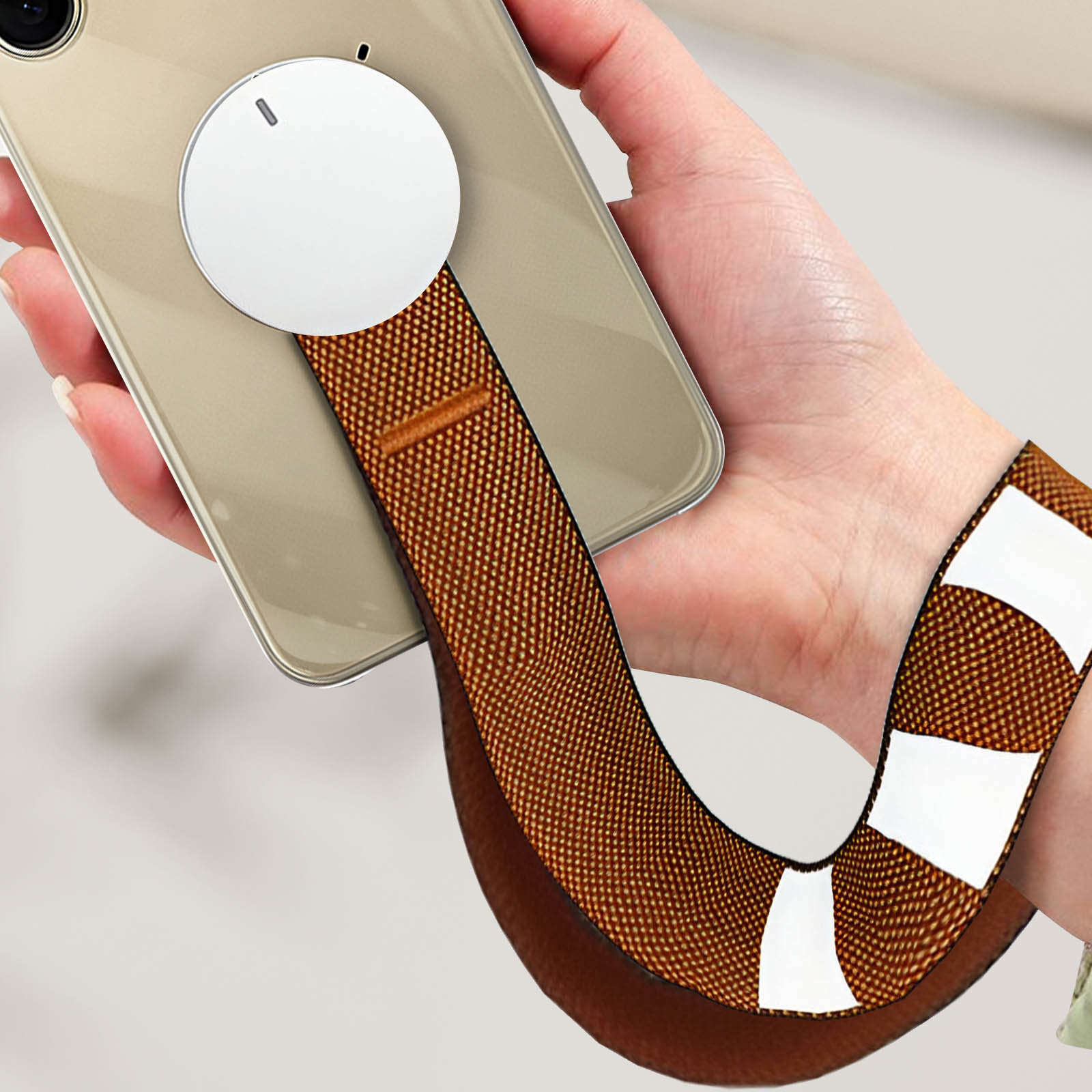 Main tenant un téléphone avec un accessoire MagSafe circulaire blanc. Sangle marron.