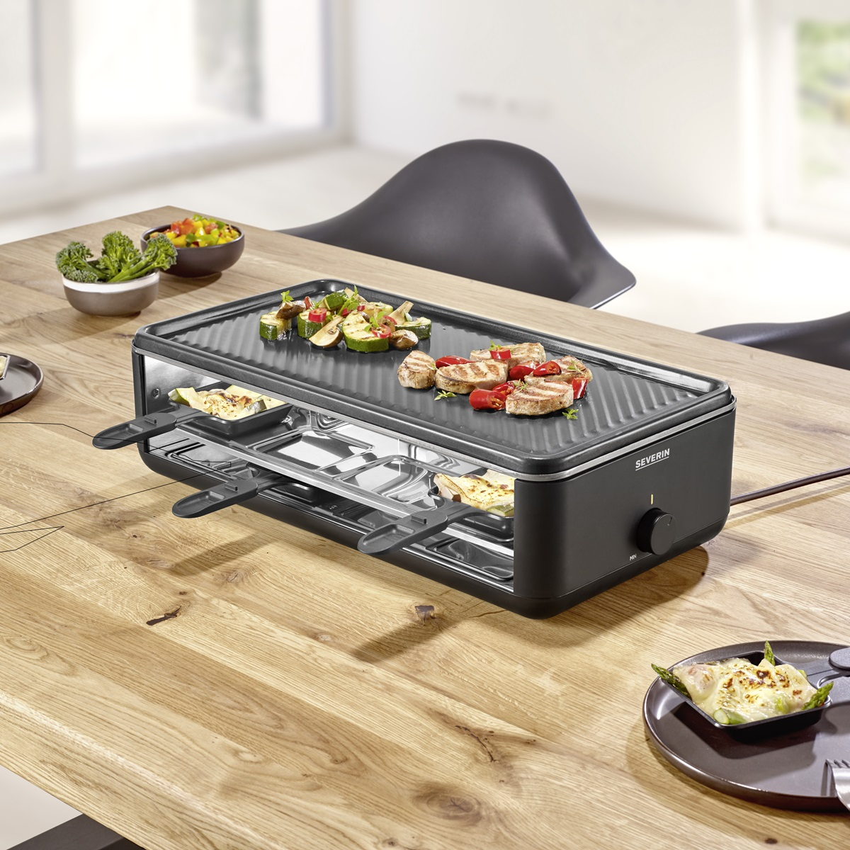 Czarny grill do raclette z jedzeniem na nim, na drewnianym stole. Brokuły i papryka w miskach.