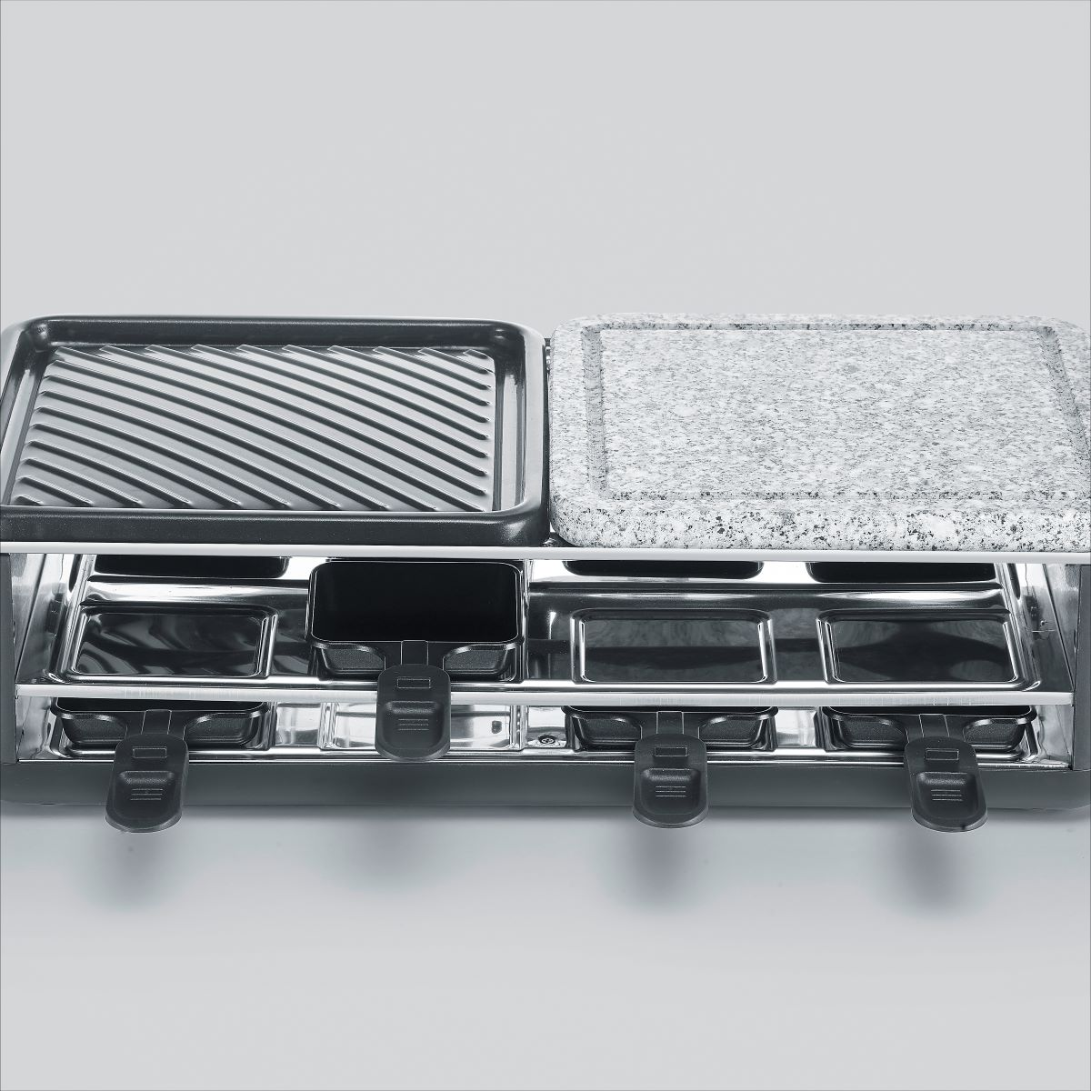 Grill do raclette. Widoczny grill, kamień i patelnie do sera. Czarne uchwyty.