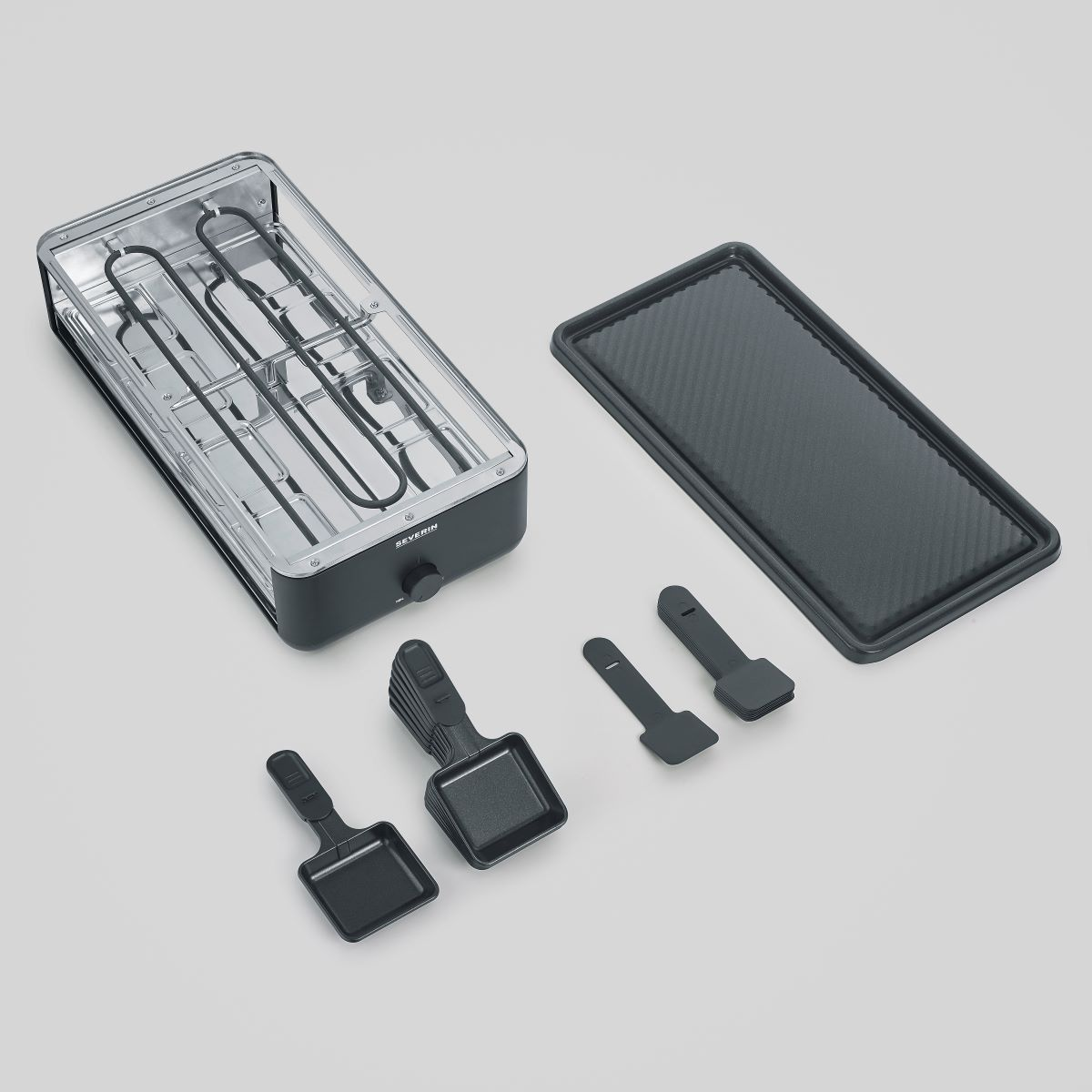 Widok z góry na grill do raclette z akcesoriami. Czarne i przezroczyste elementy na białej powierzchni.