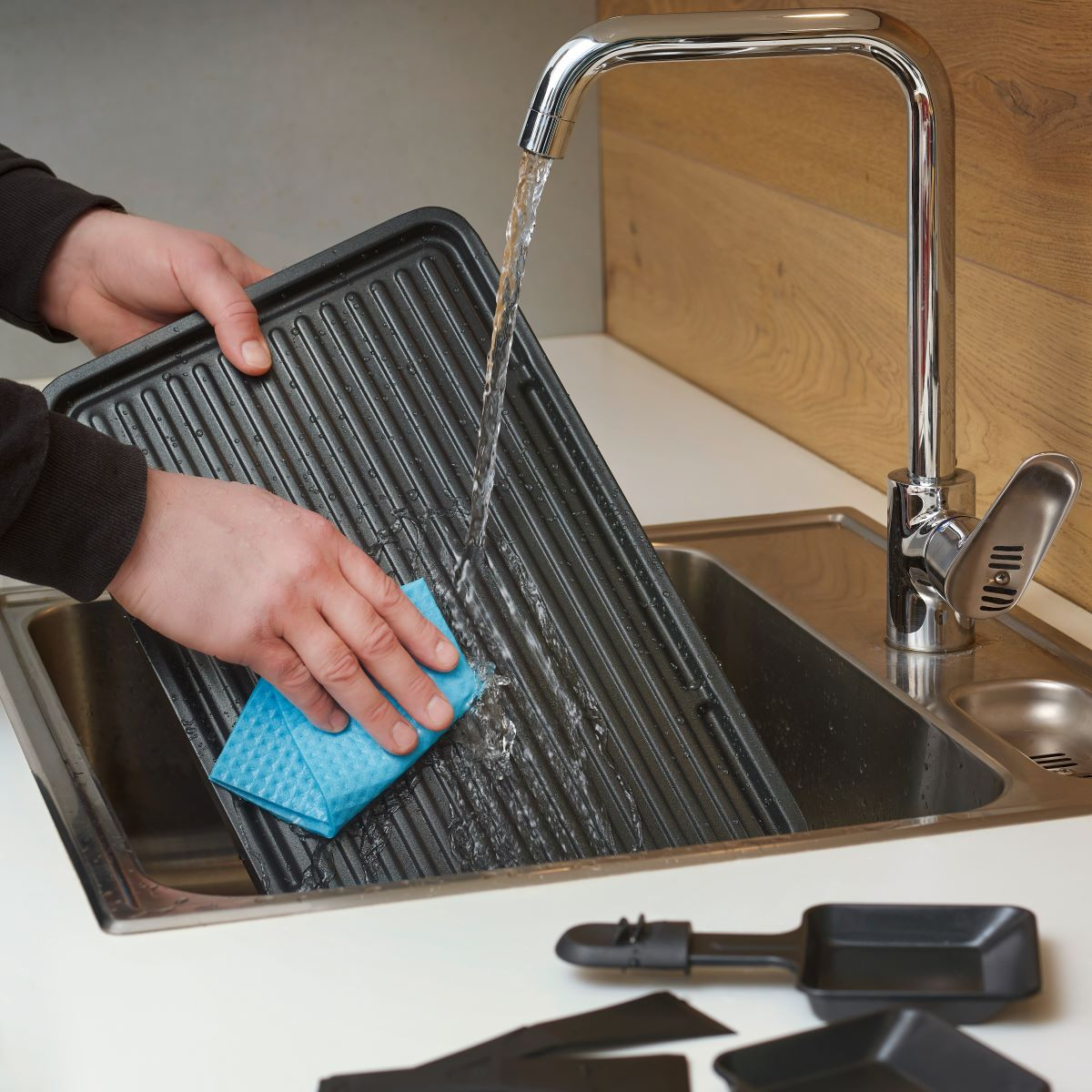 Osoba myje czarną patelnię grillową w zlewie ze stali nierdzewnej za pomocą niebieskiej gąbki i wody.