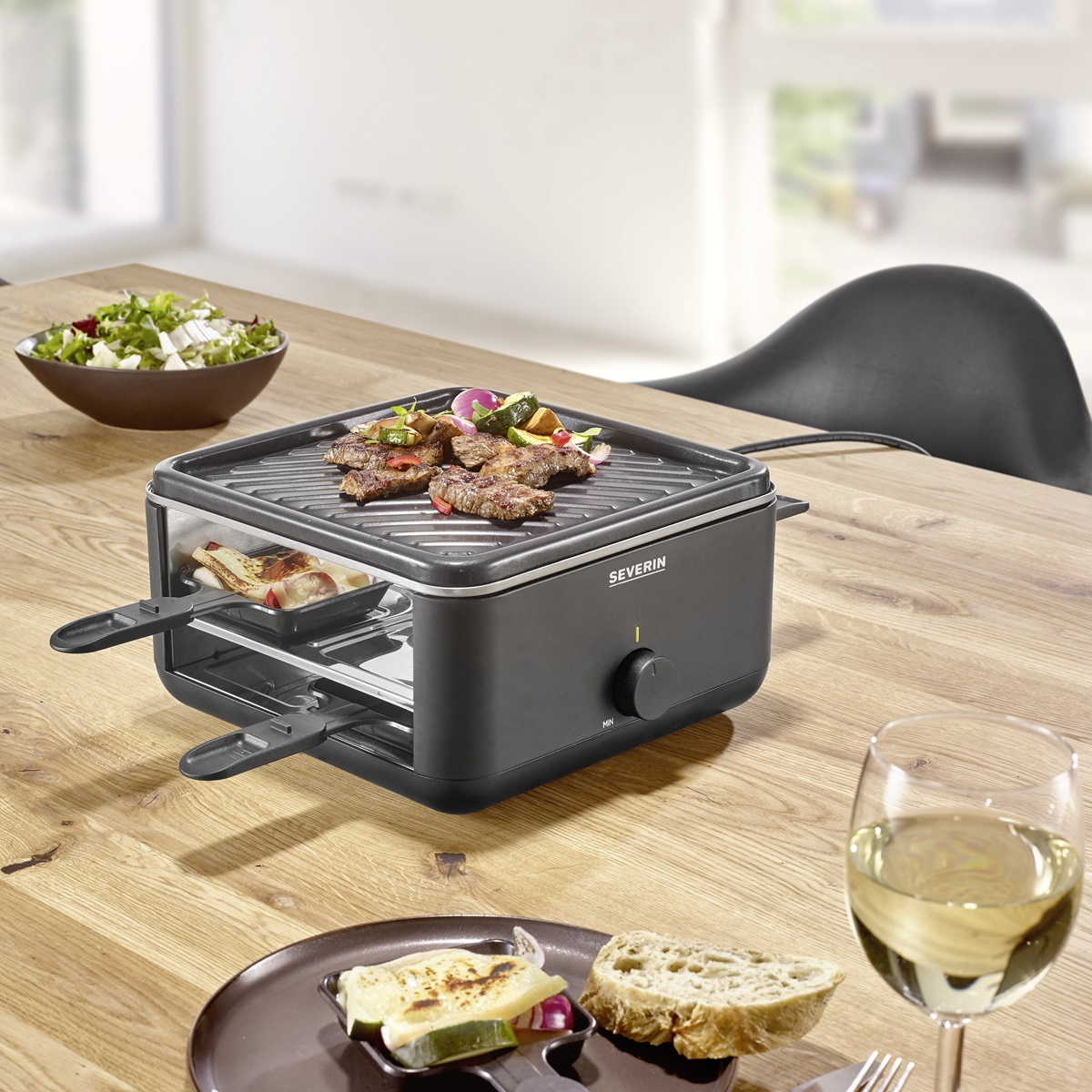 Grill do raclette na drewnianym stole, z jedzeniem. Obecny kieliszek wina i sałatka.