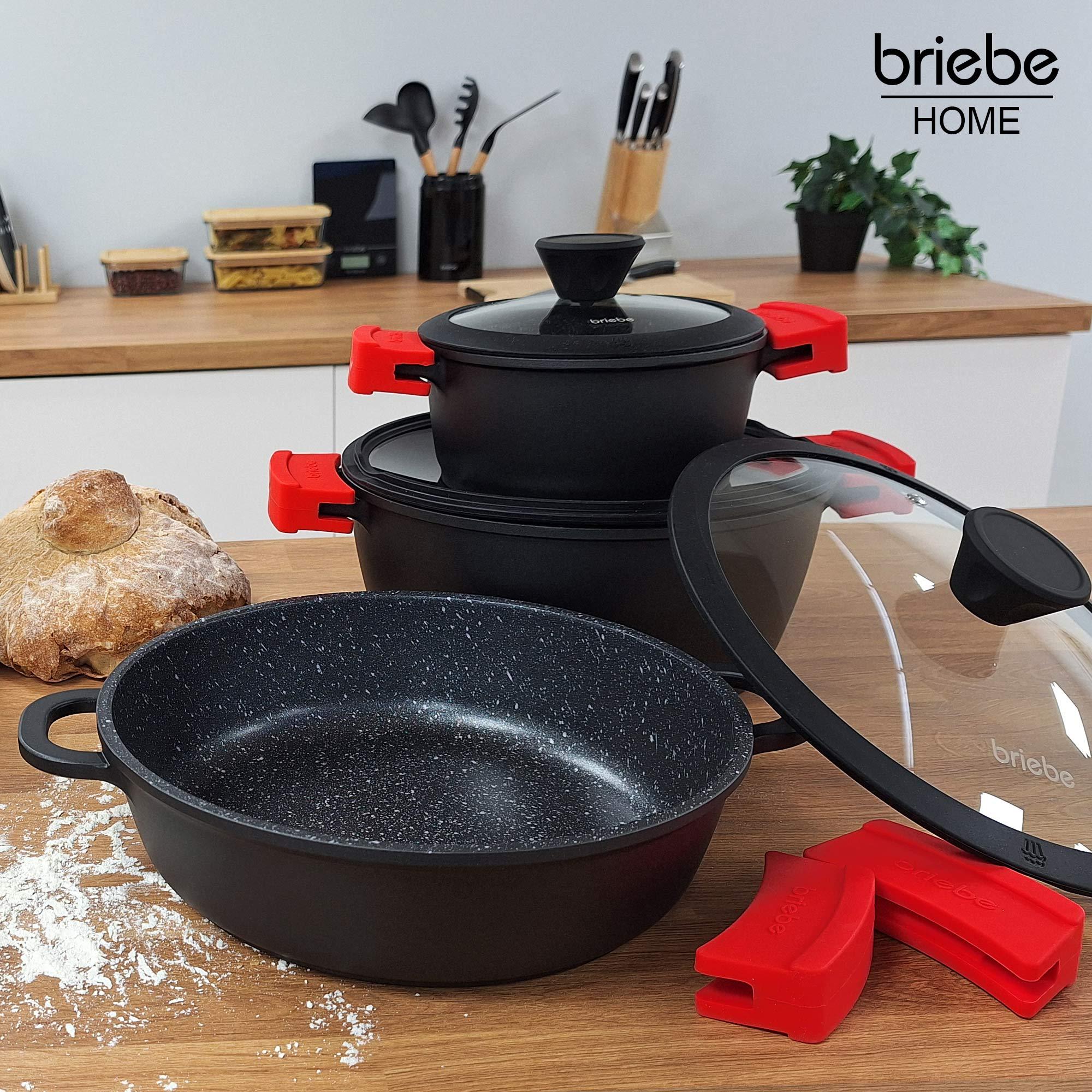 Un juego de ollas negras con asas rojas y tapas de vidrio, con varios utensilios de cocina.