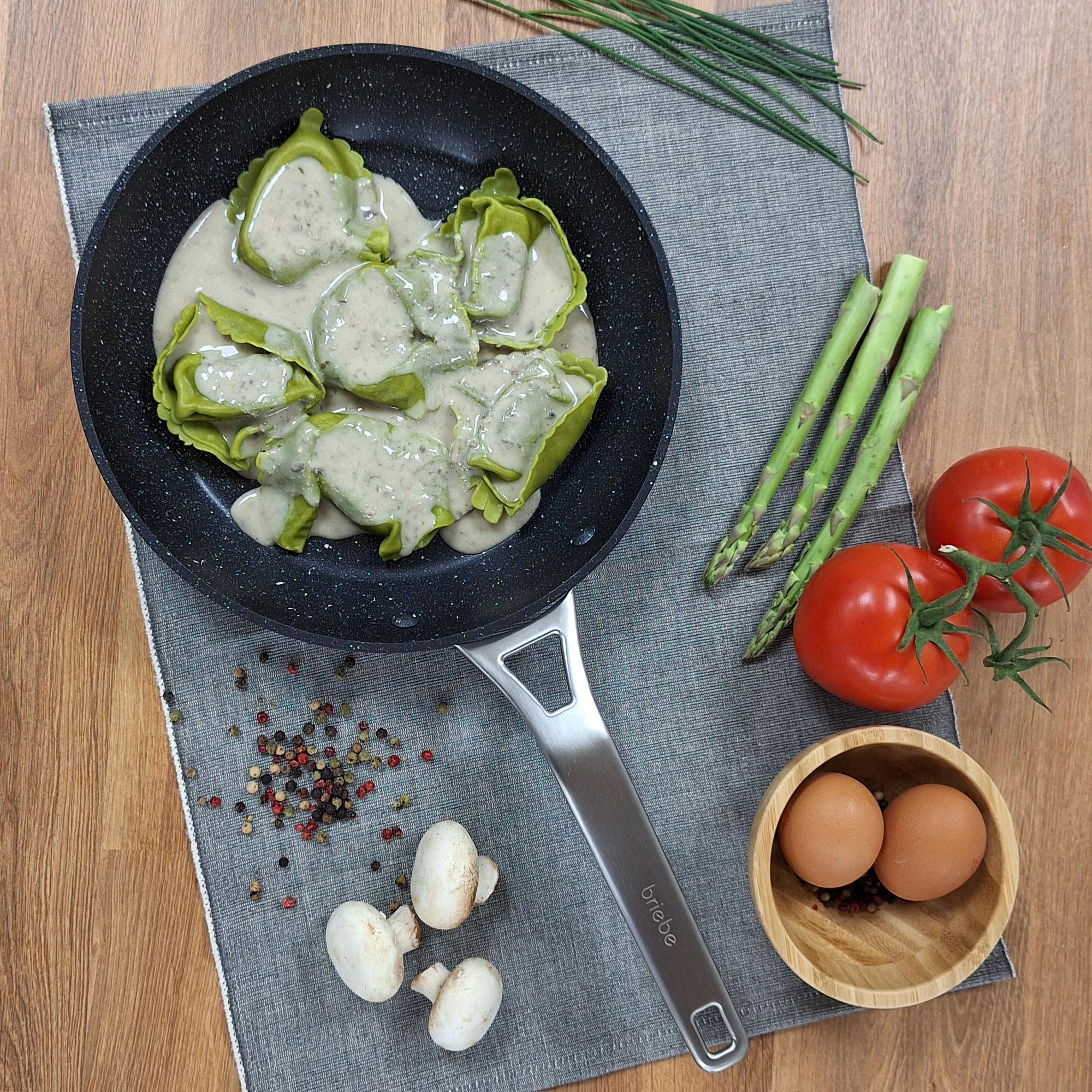 Vista cenital de una sartén con tortellini verdes en salsa, tomates, huevos, champiñones y espárragos.