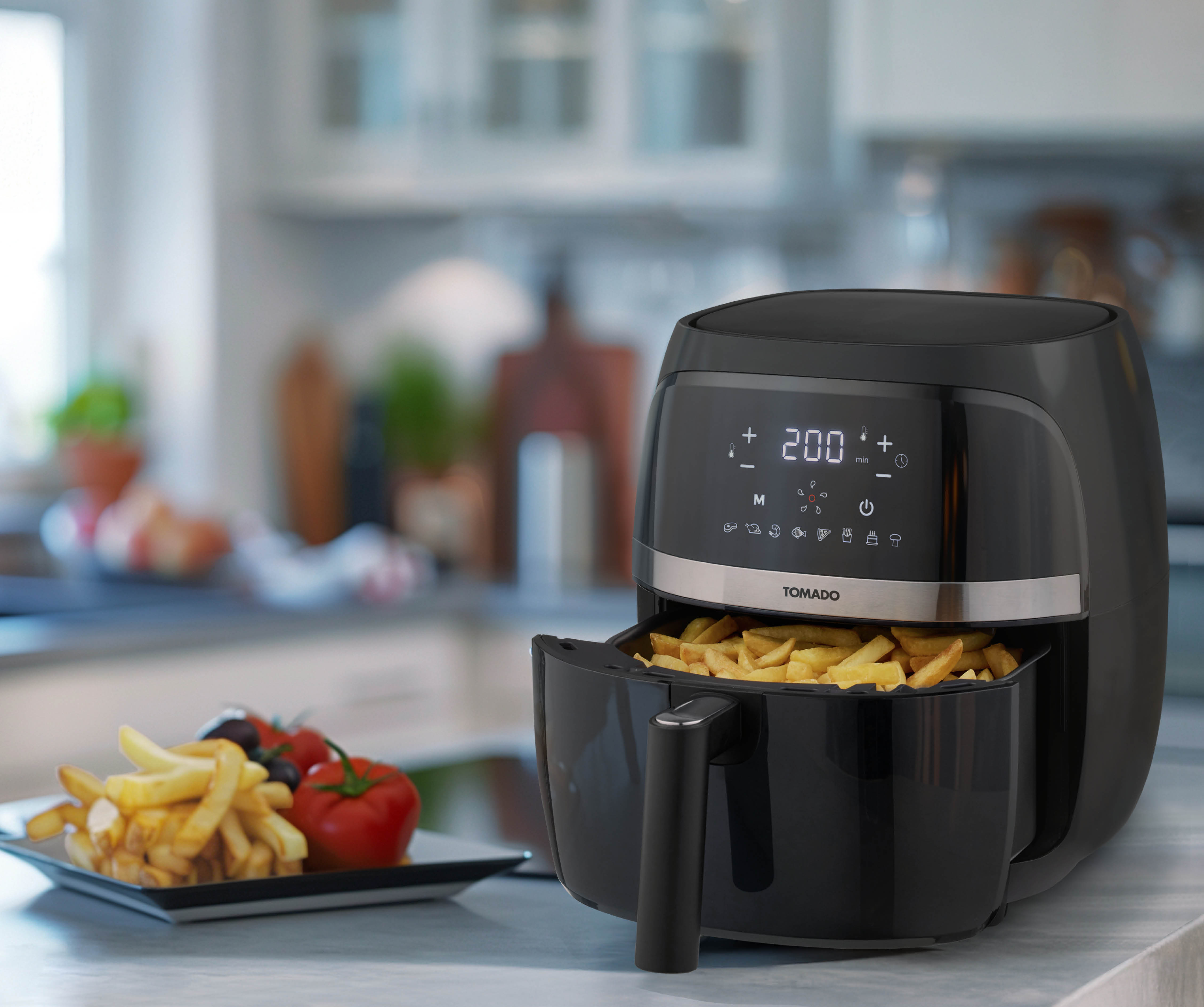 Black air fryer with fries in the basket and tomatoes on a plate on a counter.