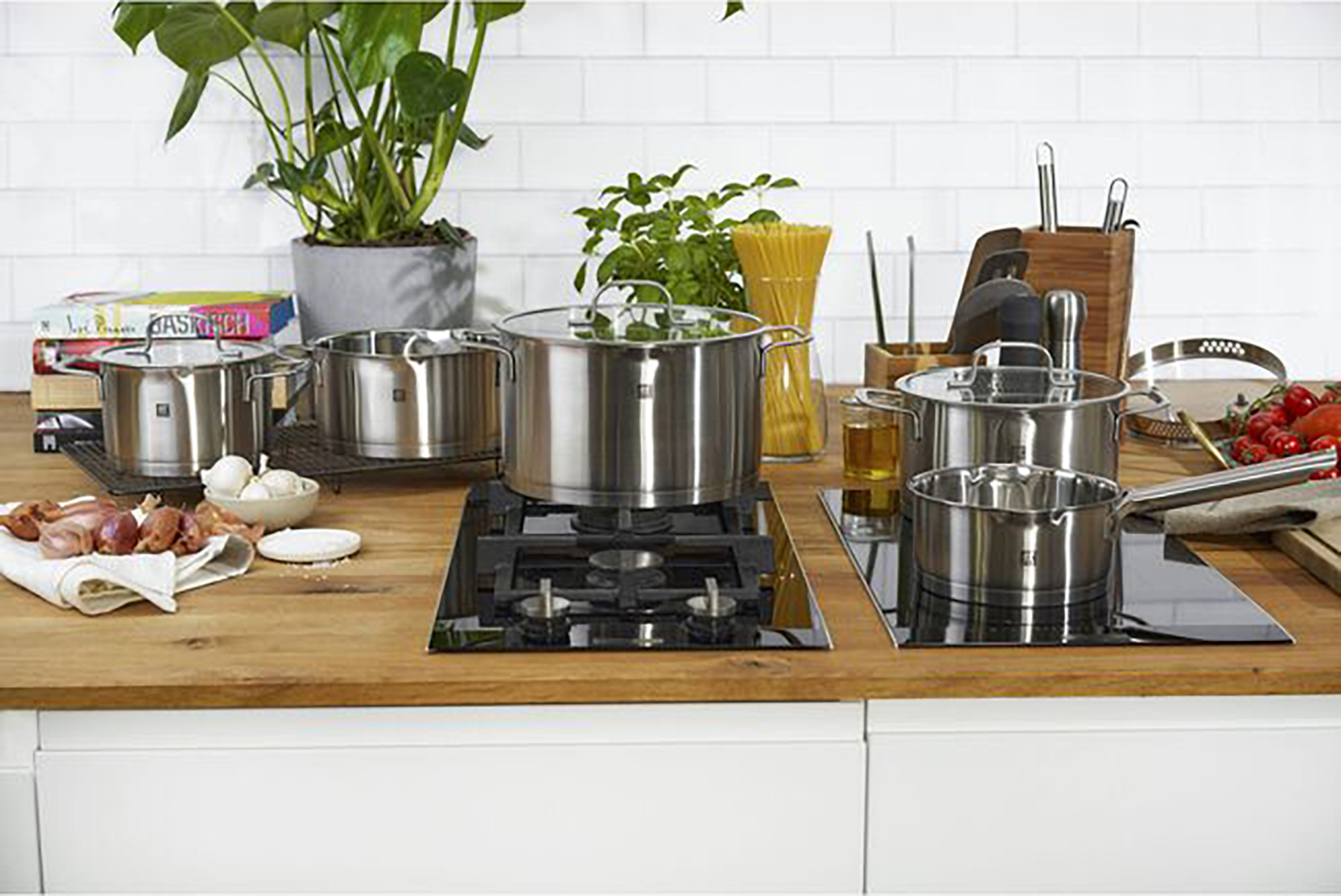 Scena kuchenna ze stalowymi garnkami i patelniami na drewnianym blacie. Biała ceglana ściana.