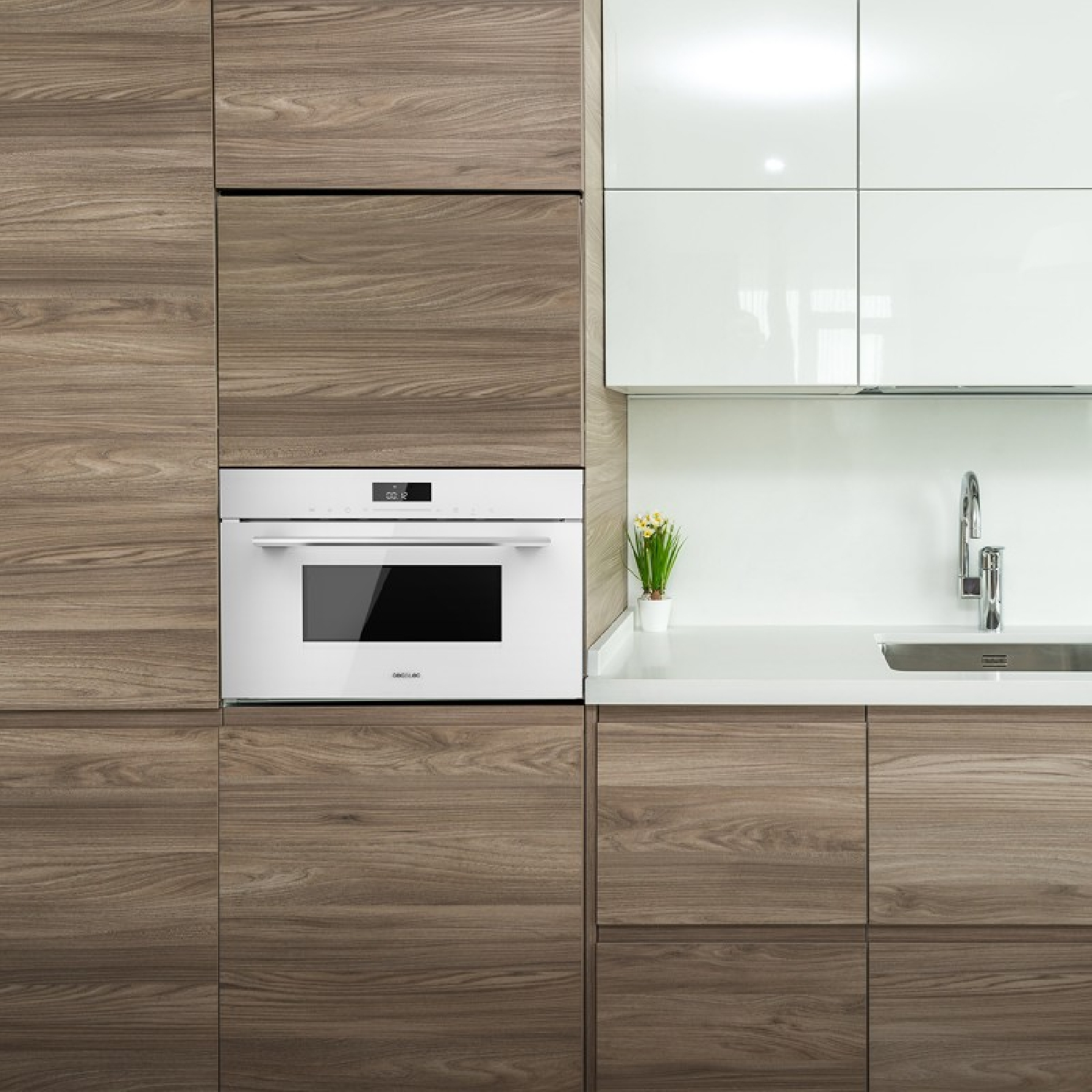 Biała kuchenka mikrofalowa wbudowana w drewnianą szafkę. W pobliżu stoi mała roślina.