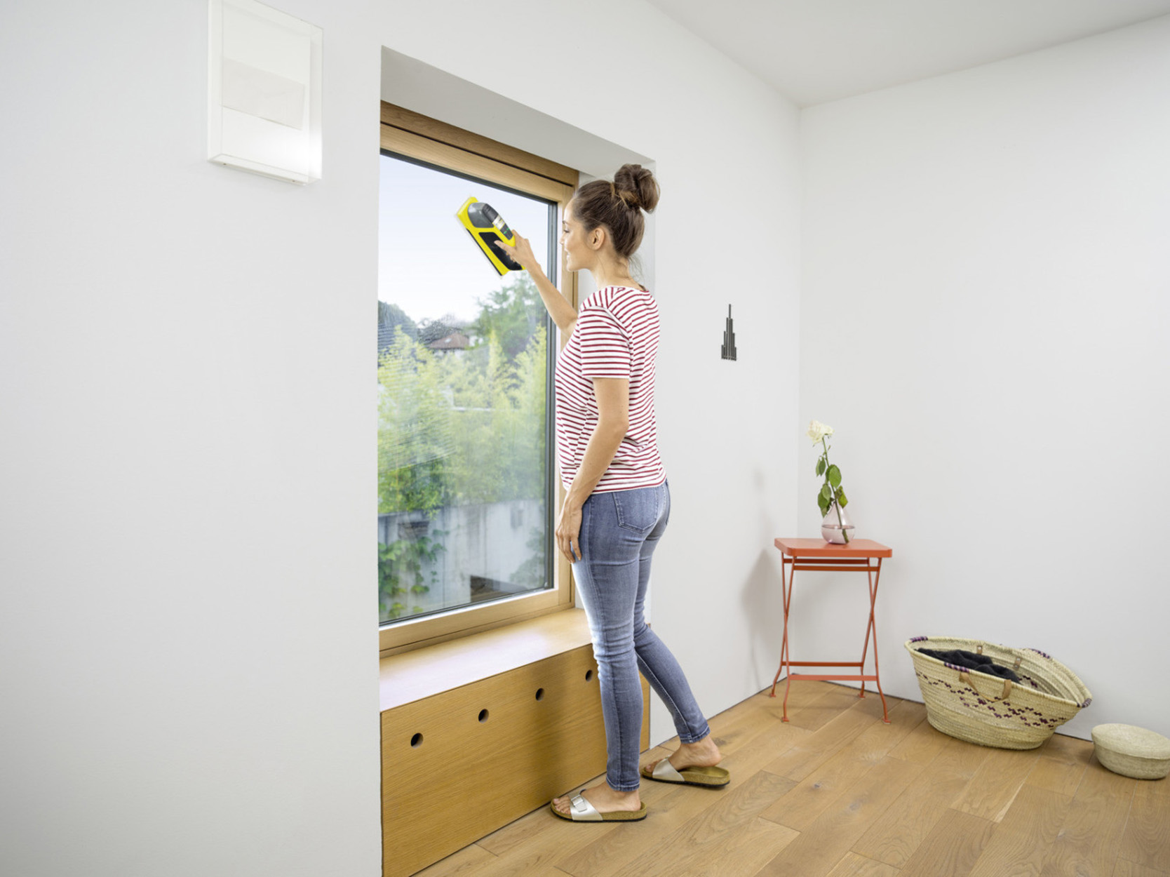 Kobieta czyszcząca okno żółtą ściągaczką. Okno znajduje się obok zielonego otoczenia.