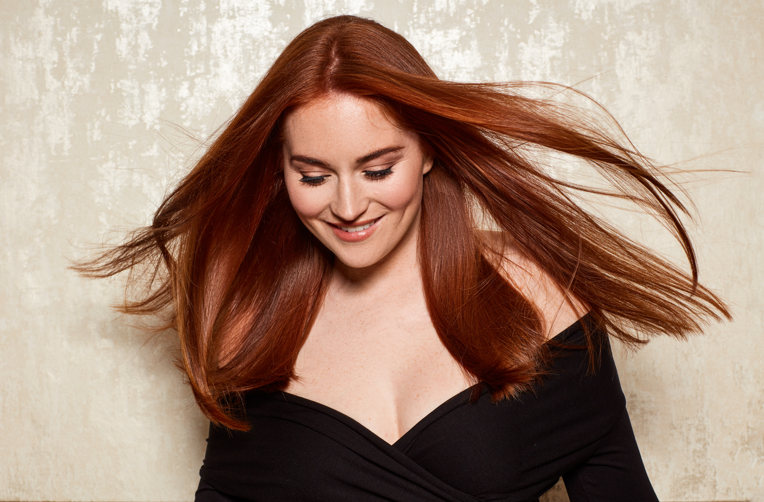 Een vrouw met lang rood haar, die naar beneden kijkt en lacht. Ze draagt een zwarte off-the-shoulder top.