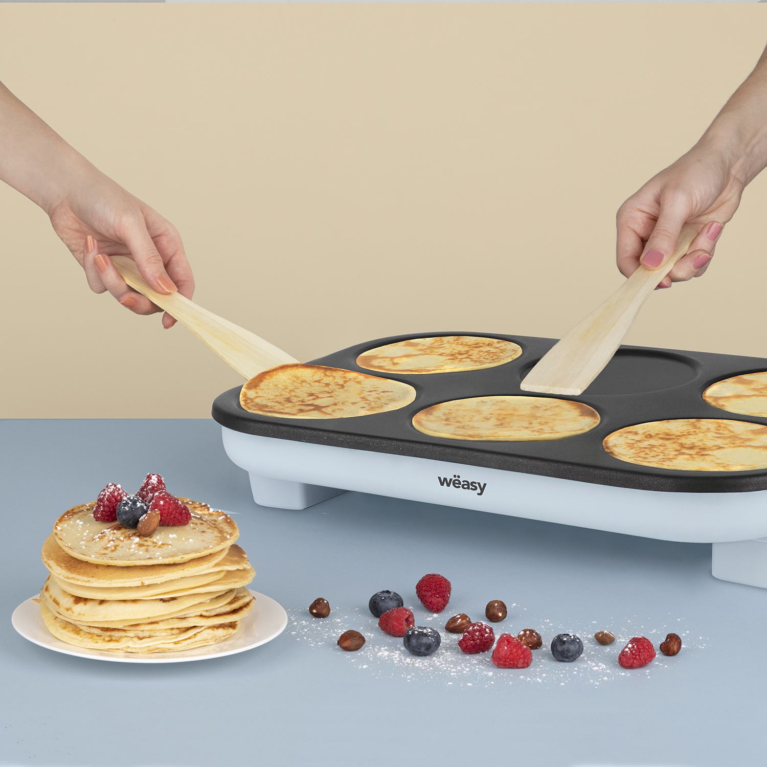 Zwei Hände wenden Pfannkuchen auf einer Grillplatte. Ein Stapel Pfannkuchen und Beeren auf einem Teller. Beeren und Nüsse sind verstreut.