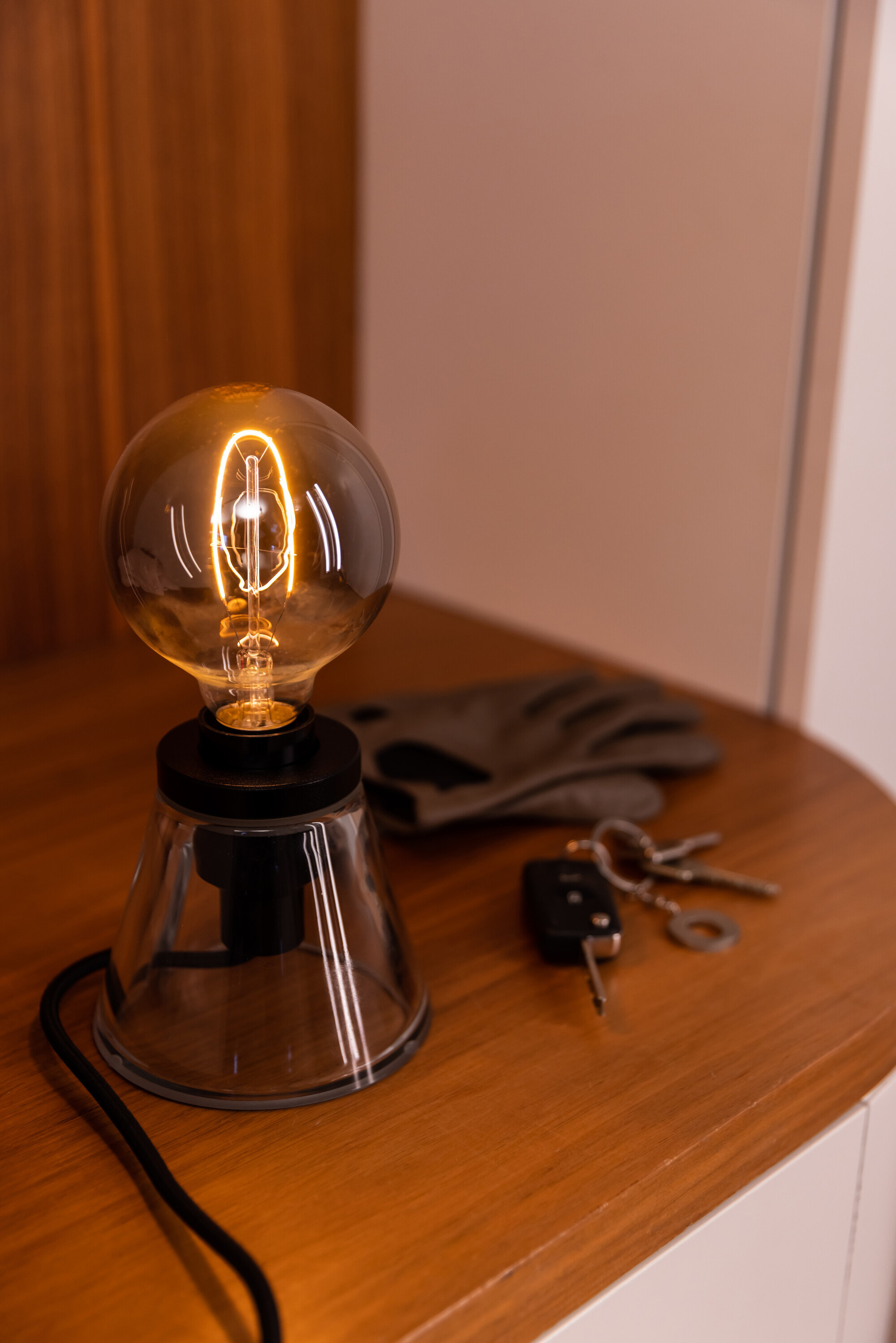 Lampe à ampoule allumée sur une surface en bois. Un gant et des clés à côté. Fond blanc.