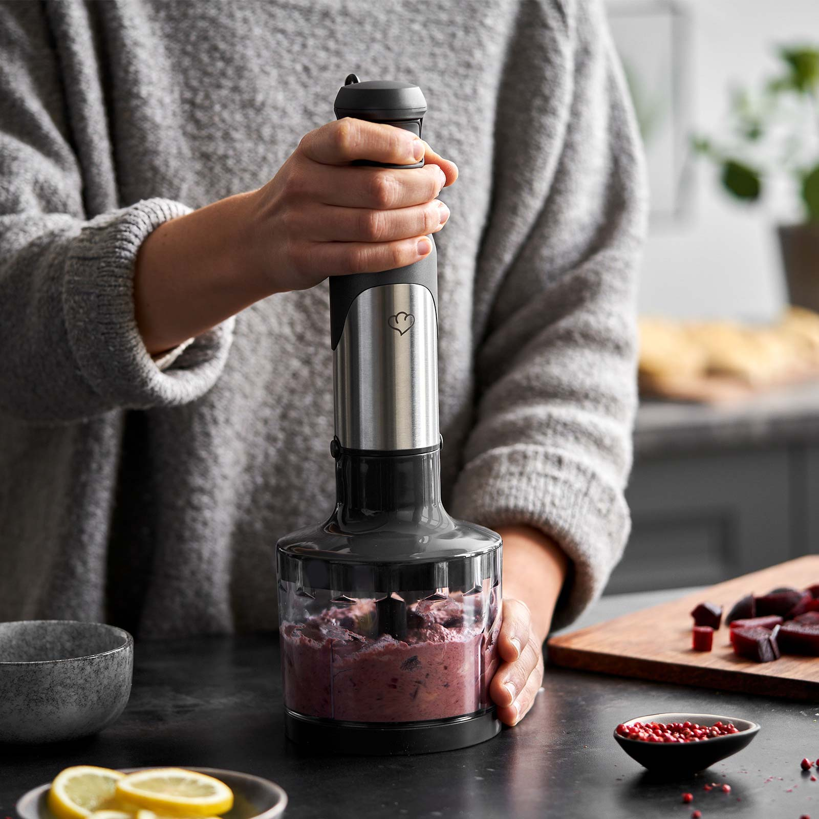 Person verrührt Essen mit einem Stabmixer. Zutaten, eine Schüssel mit Zitrone und ein Schneidebrett sind sichtbar.