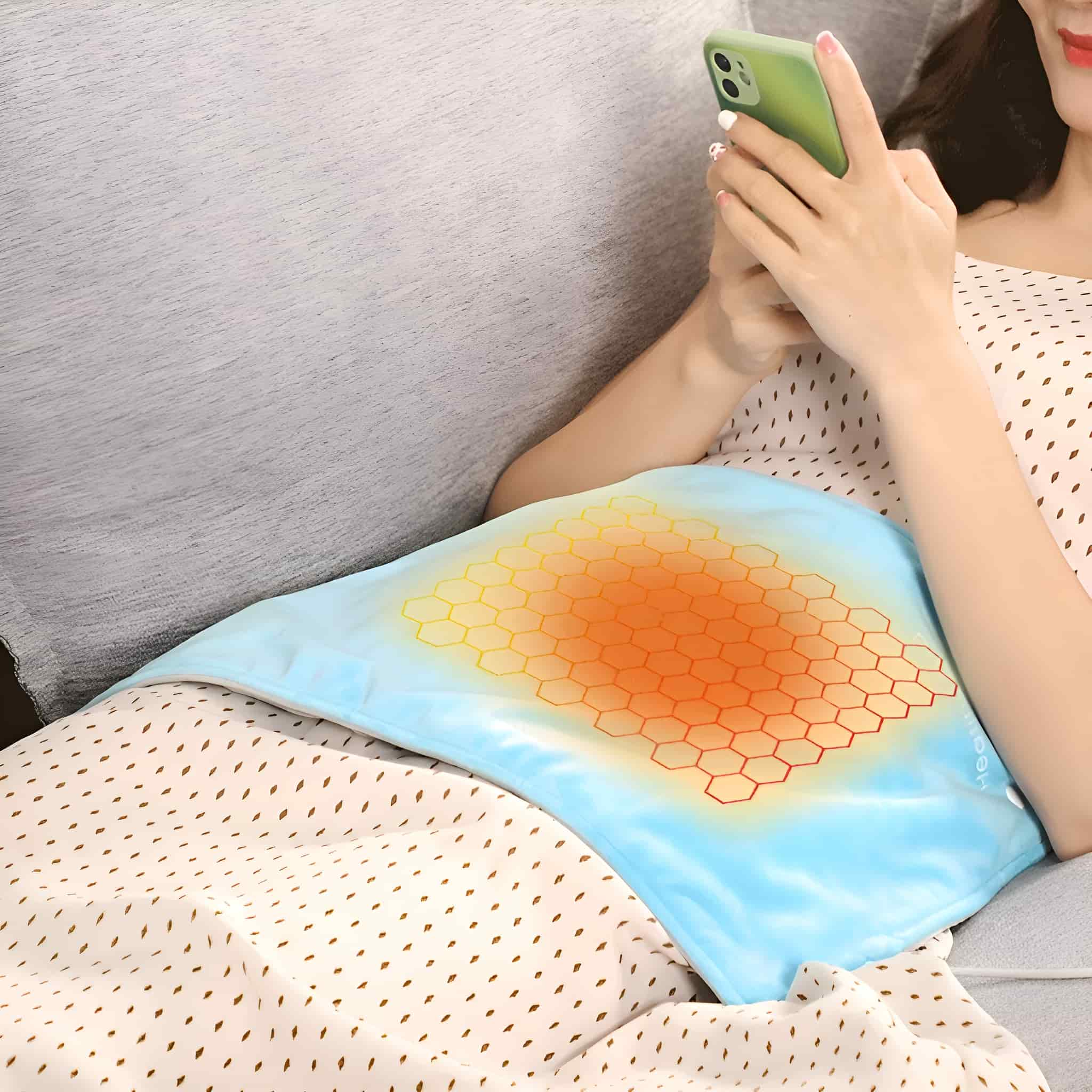 Eine Frau auf einem Sofa mit einem blauen Heizkissen auf dem Schoß, die ihr Telefon benutzt. Das Kissen ist beleuchtet.