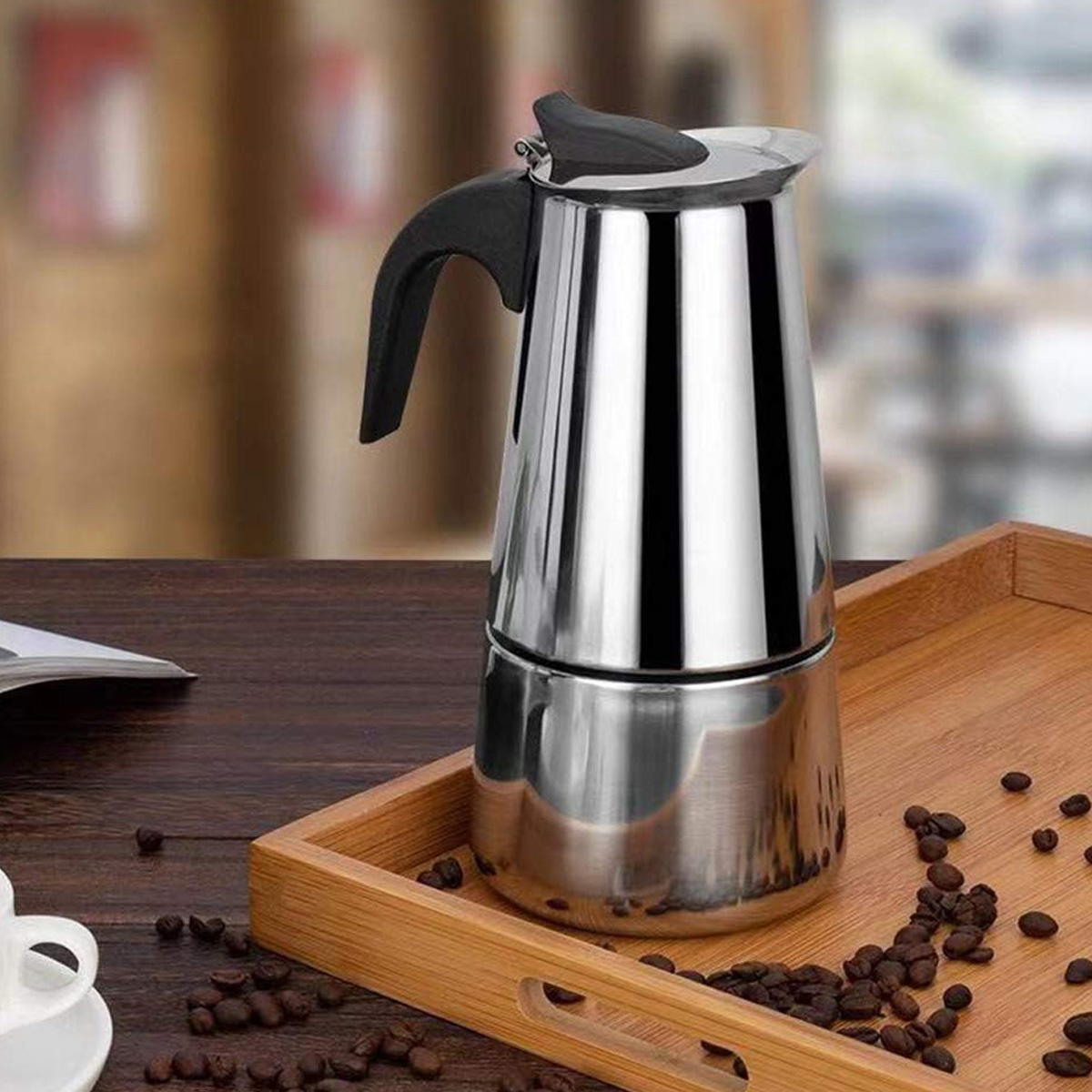 Glänzender silberner Kaffeezubereiter auf einem Holztablett mit Kaffeebohnen. Der Hintergrund ist unscharf.