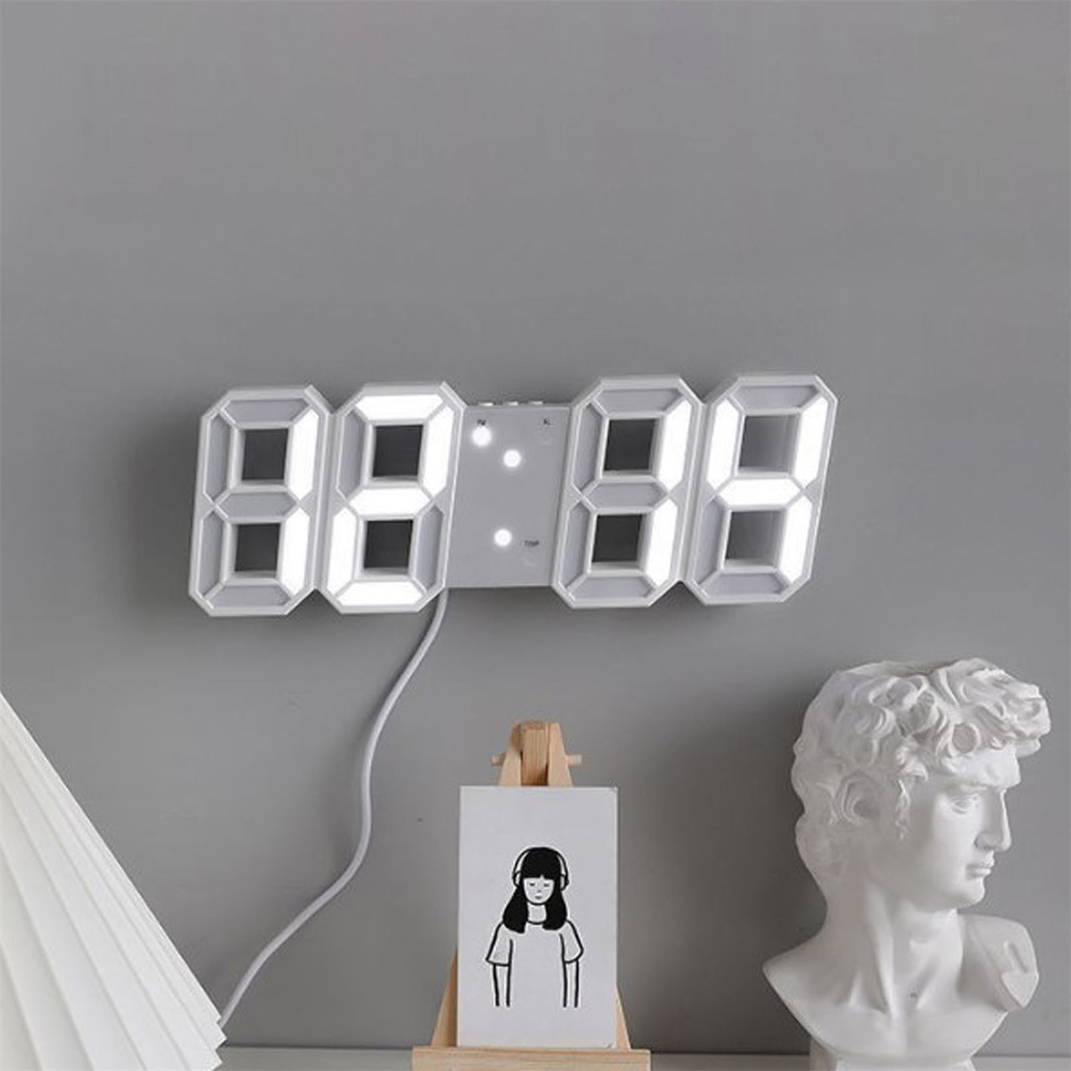 Weiße Digitaluhr an einer grauen Wand mit Büste und Kunstwerk darunter.