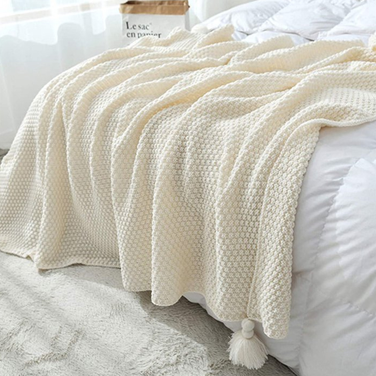 Cremefarbene Strickdecke mit Quasten auf einem Bett mit weißen Laken. Die Decke ist ordentlich gefaltet.