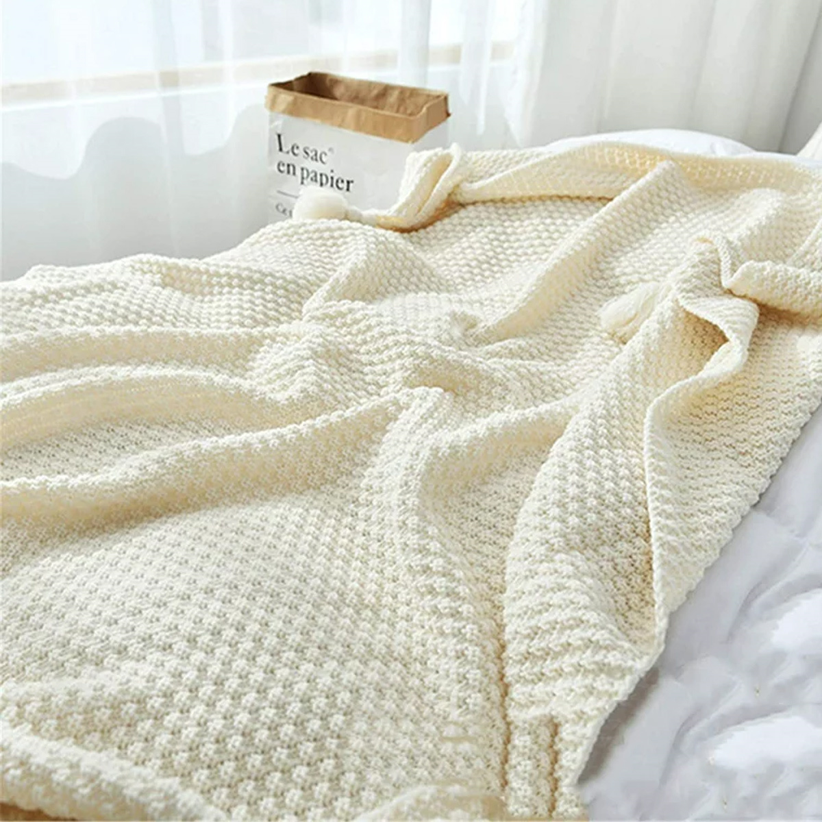 Cremefarbene Strickdecke auf einem weißen Bett, mit einer Papiertüte im Hintergrund.