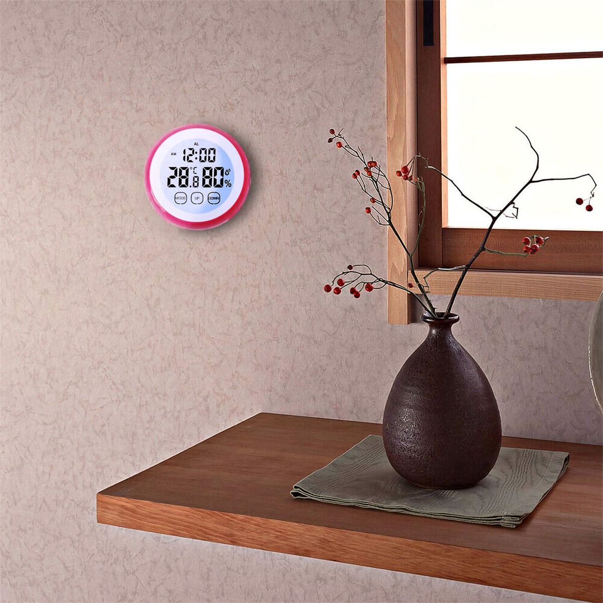Ein digitales Thermometer mit rosa Rahmen zeigt Temperatur und Luftfeuchtigkeit neben einer Vase und Fenster.