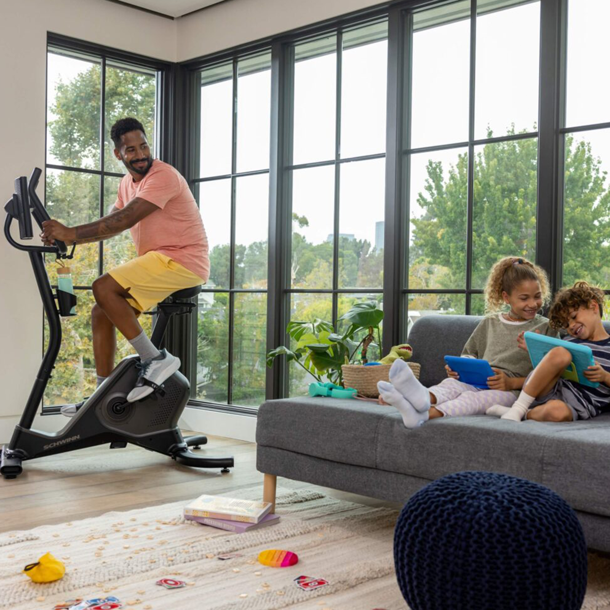 Un homme sur un vélo d'appartement et deux enfants sur un canapé utilisant des tablettes avec une grande fenêtre en arrière-plan.