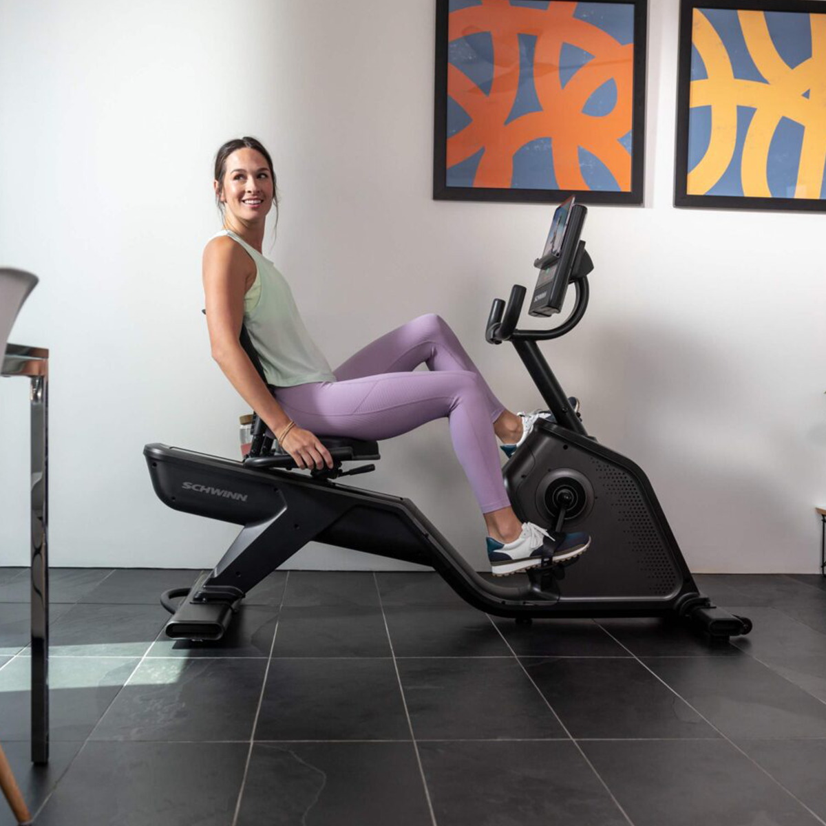 Frau auf einem Liege-Heimtrainer. Sie schaut in die Kamera, heller Raum mit Kunst an der Wand.