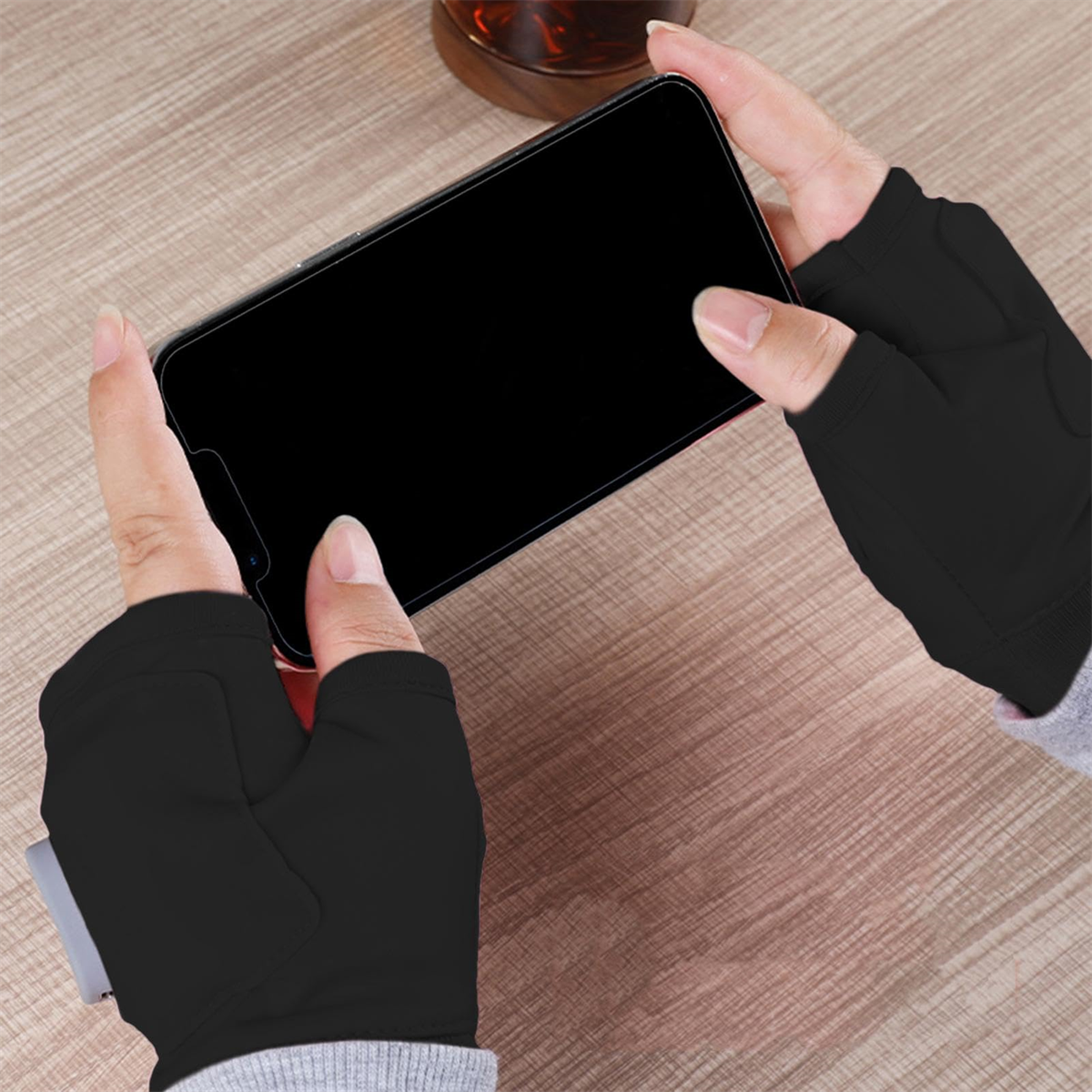 Hände einer Person mit schwarzen Handschuhen, die ein Telefon mit schwarzem Bildschirm auf einem Holztisch halten.