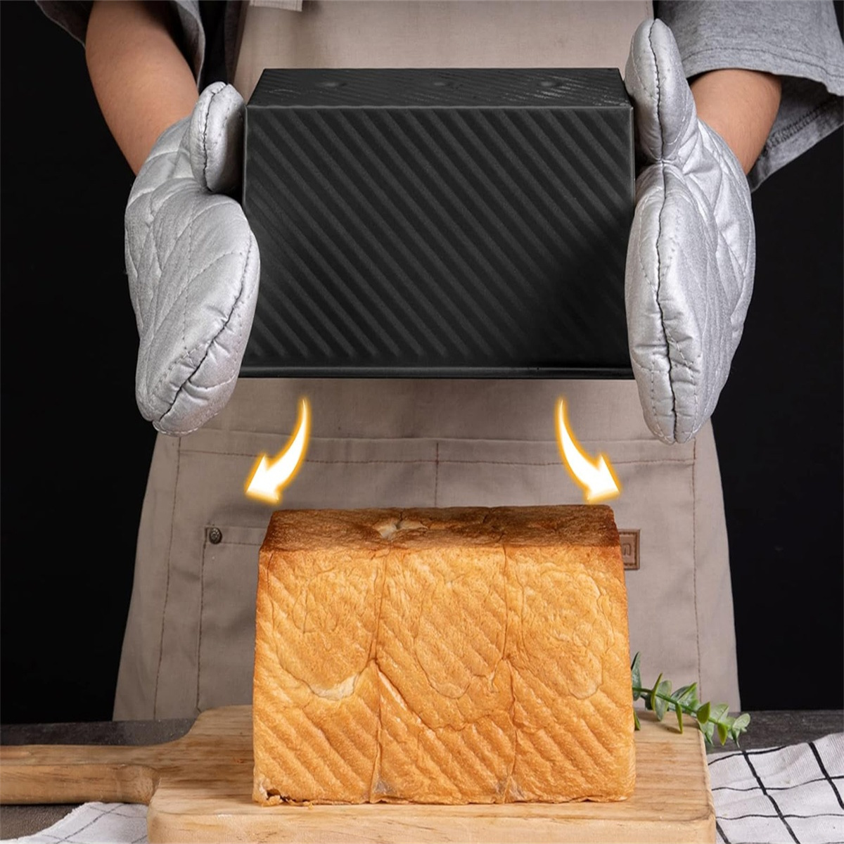 Ein Brotlaib wird mit Ofenhandschuhen aus einer schwarzen Brotform genommen. Holzbrett und Schürze vorhanden.