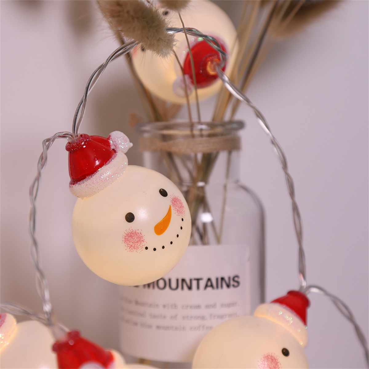 Weihnachtsbeleuchtung mit Schneemann-Ornamenten und roten Weihnachtsmützen, vor einer Vase und Trockenblumen.