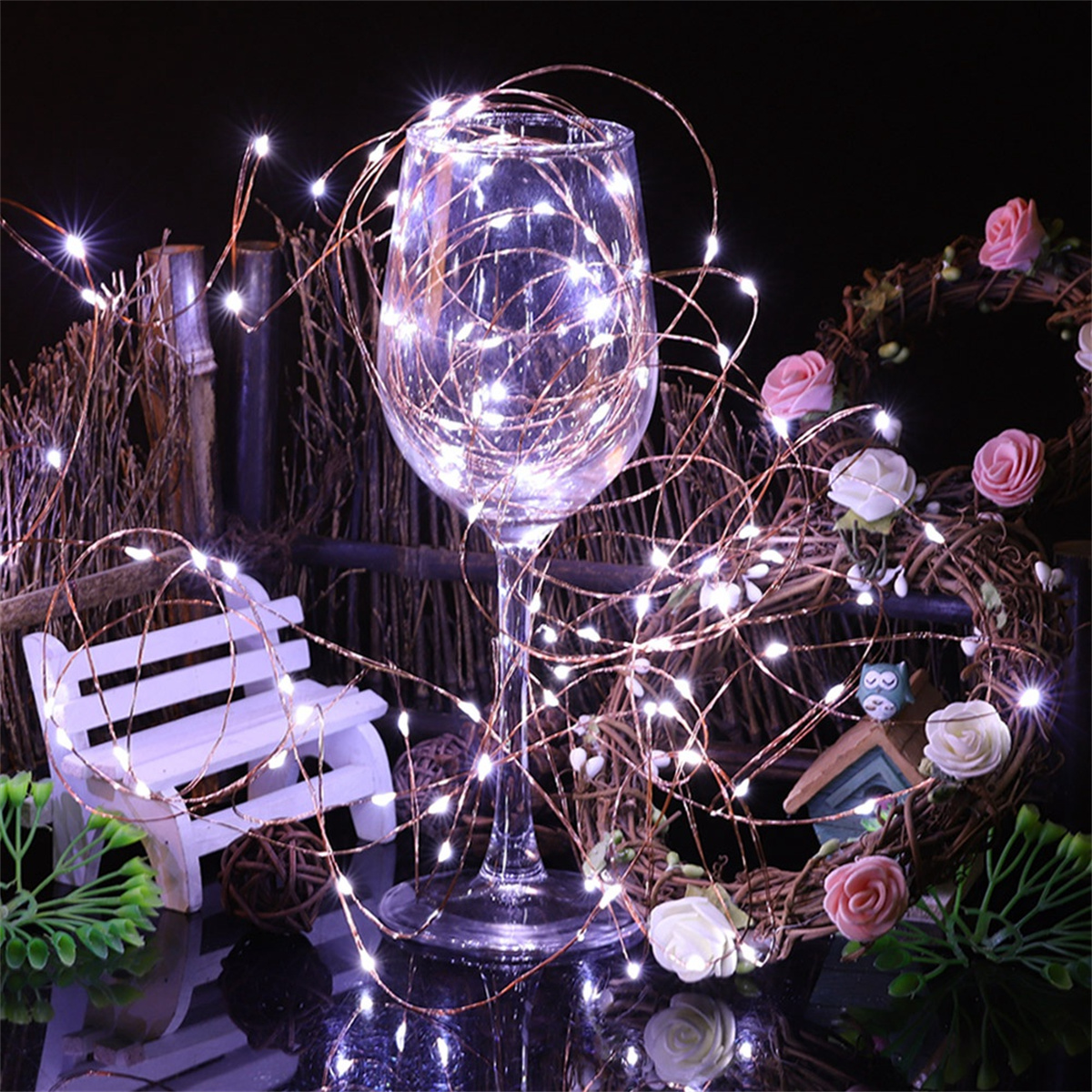 Ein Glas gefüllt mit Lichterketten. Rosen, eine kleine Bank und ein Kranz umgeben es. Dunkler Hintergrund.
