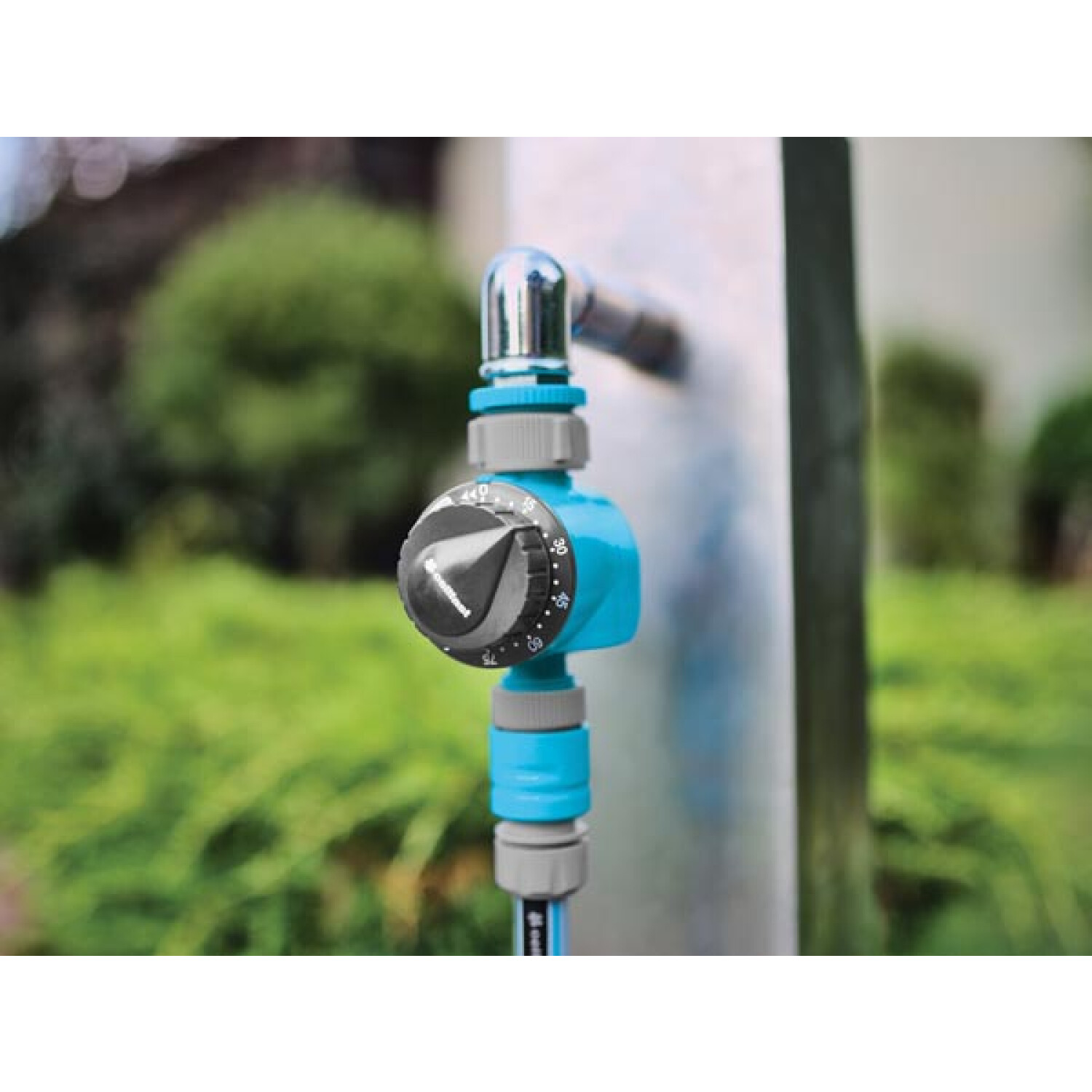 Blauer und grauer Wasserzeitschalter an einem Metallrohr, mit grünem Laub im Hintergrund.