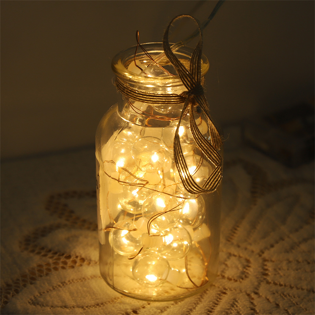 Ein klares Glasgefäß mit warmweißen Lichtern im Inneren, gebunden mit einem braunen Band. Der Hintergrund ist unscharf.