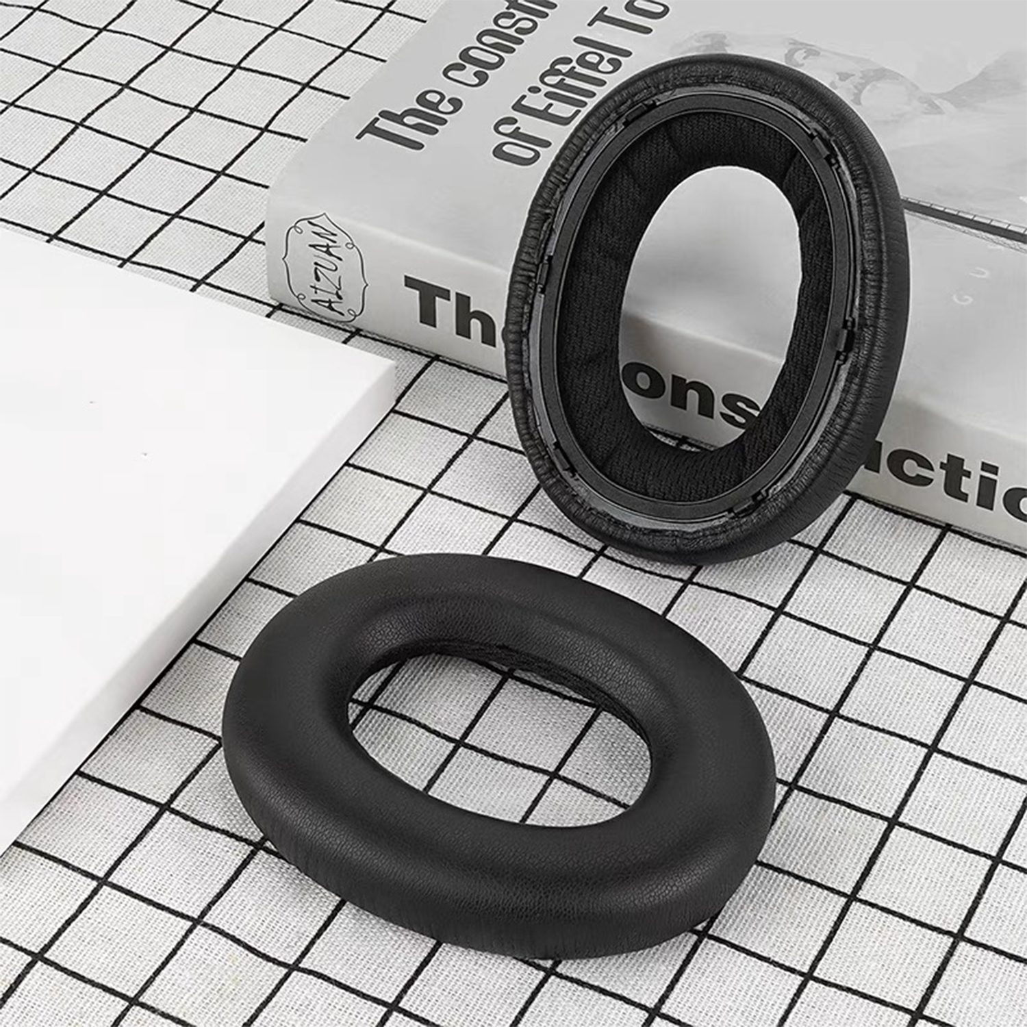 Dwie czarne poduszki nauszne do słuchawek; jedna otwarta, druga zamknięta. W tle leży książka.