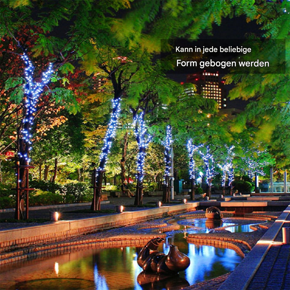 Stadtpark bei Nacht. Bäume mit blauen Lichtern. Wasser mit Statuen. Gebäude im Hintergrund.
