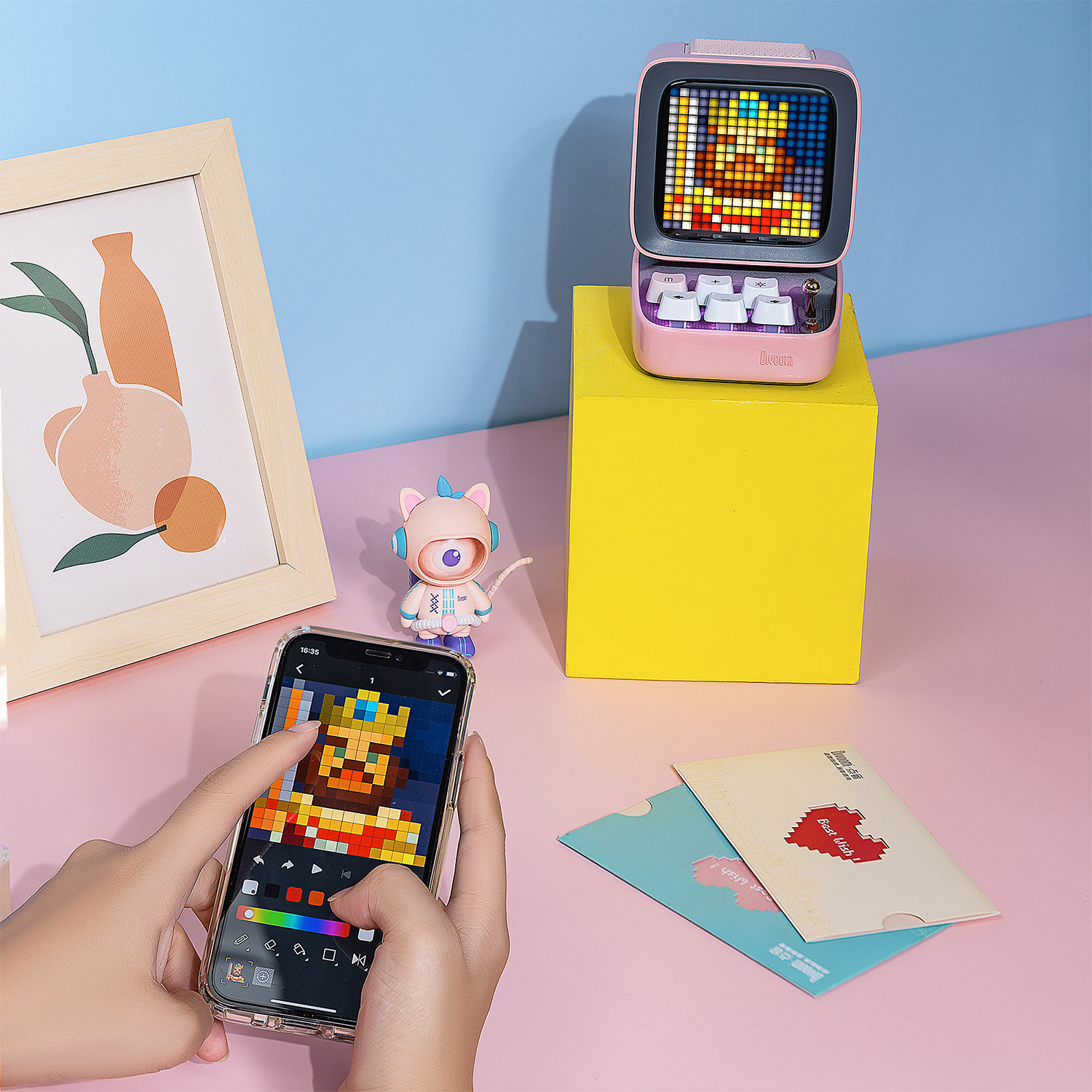 Piksel sanatı gösteren bir ekranı olan pembe bir hoparlör. Bir kişi piksel sanatı olan bir telefon kullanıyor.