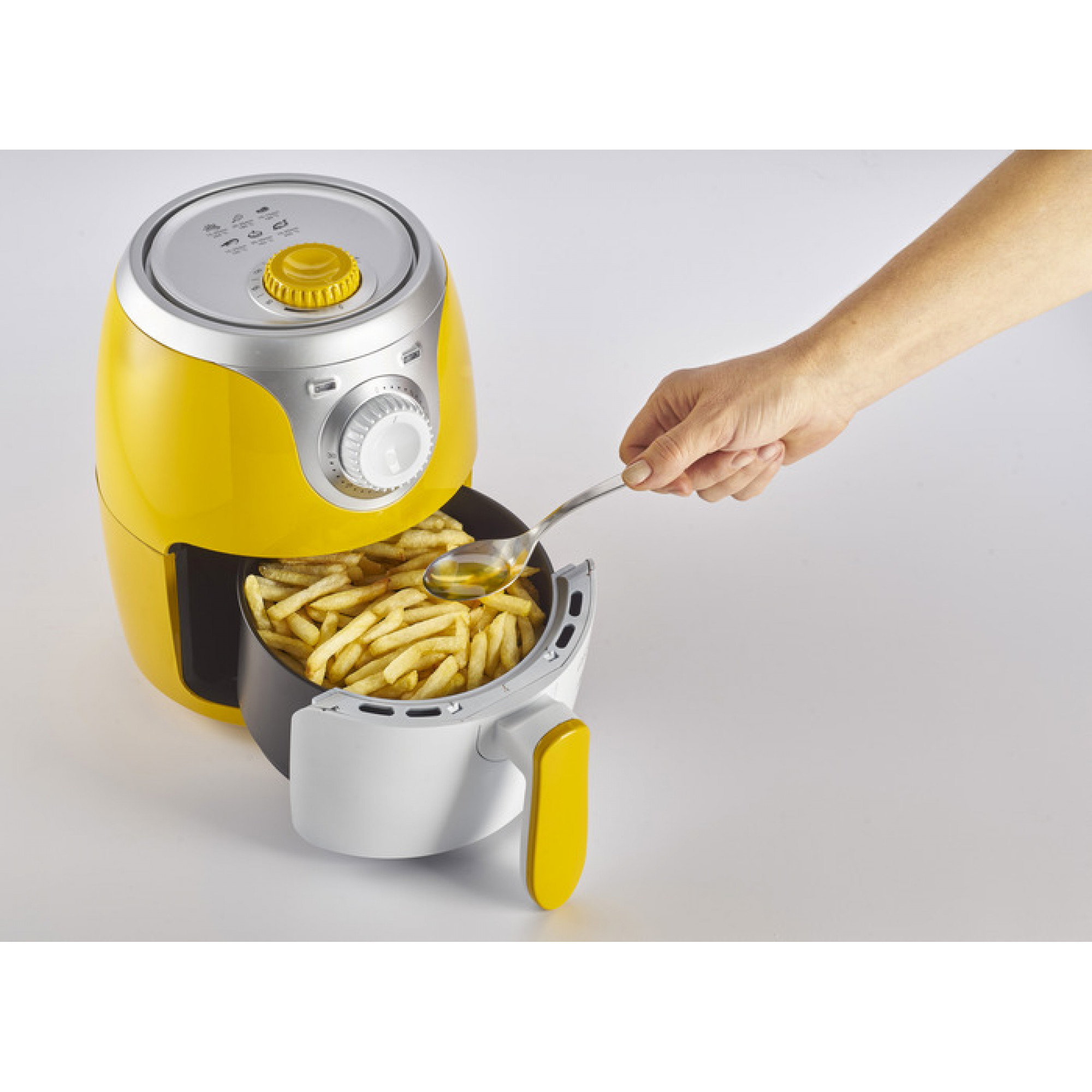 Freidora de aire amarilla con papas fritas. Una mano vierte aceite en la canasta con una cuchara.