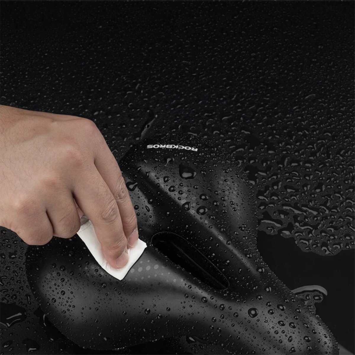 Ręka czyszcząca czarne siodełko rowerowe białą szmatką. Widoczne krople wody.