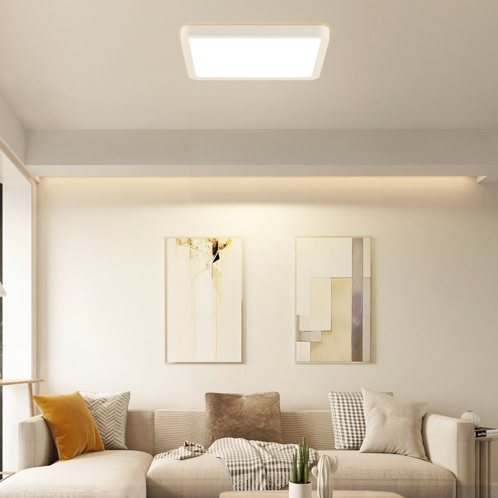 Kwadratowa biała lampa sufitowa w salonie. Widoczne dwa obrazy, sofa, poduszki i roślina.