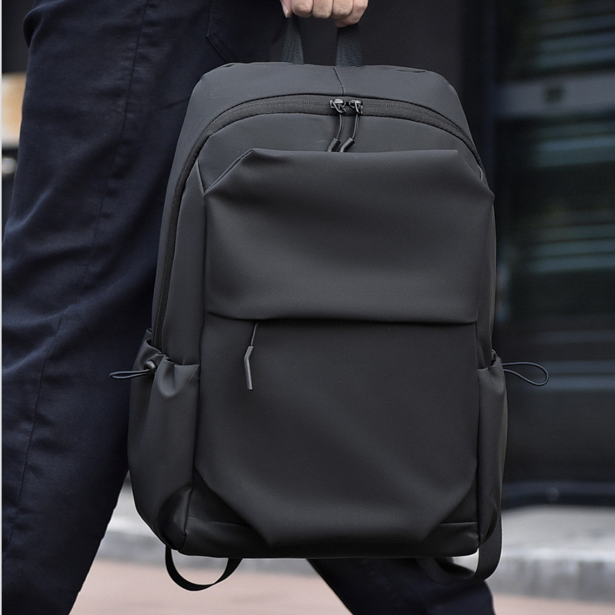 Eine Person trägt einen schwarzen Rucksack mit gepolsterten Trägern, einer Vordertasche und Seitentaschen.