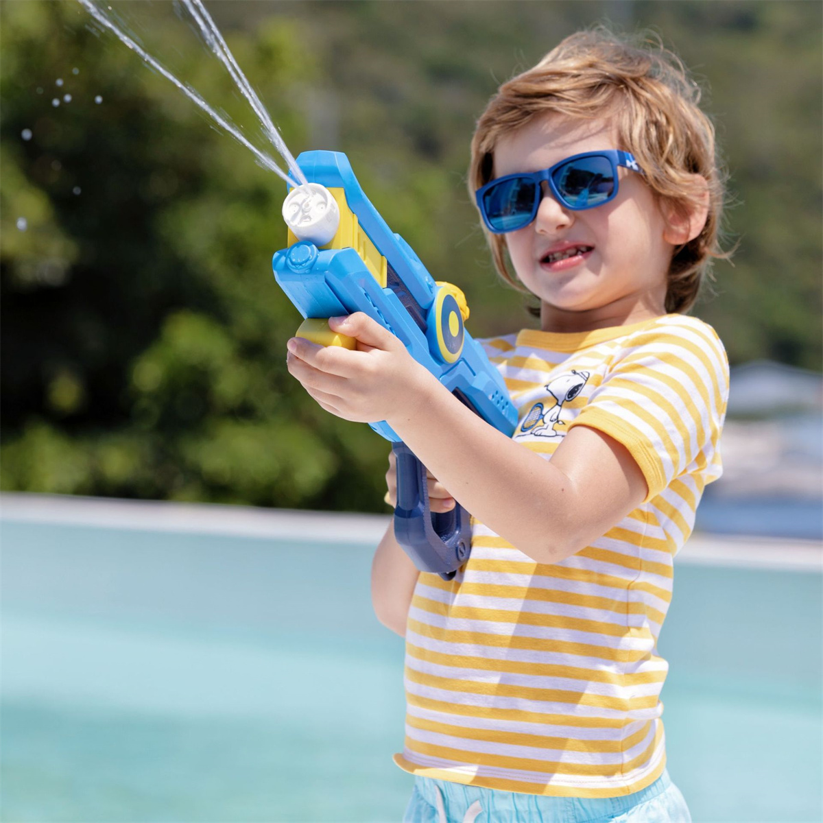 Ein kleiner Junge in Sonnenbrille spritzt Wasser aus einer blau-gelben Spielzeug-Wasserpistole. Blauer Hintergrund.