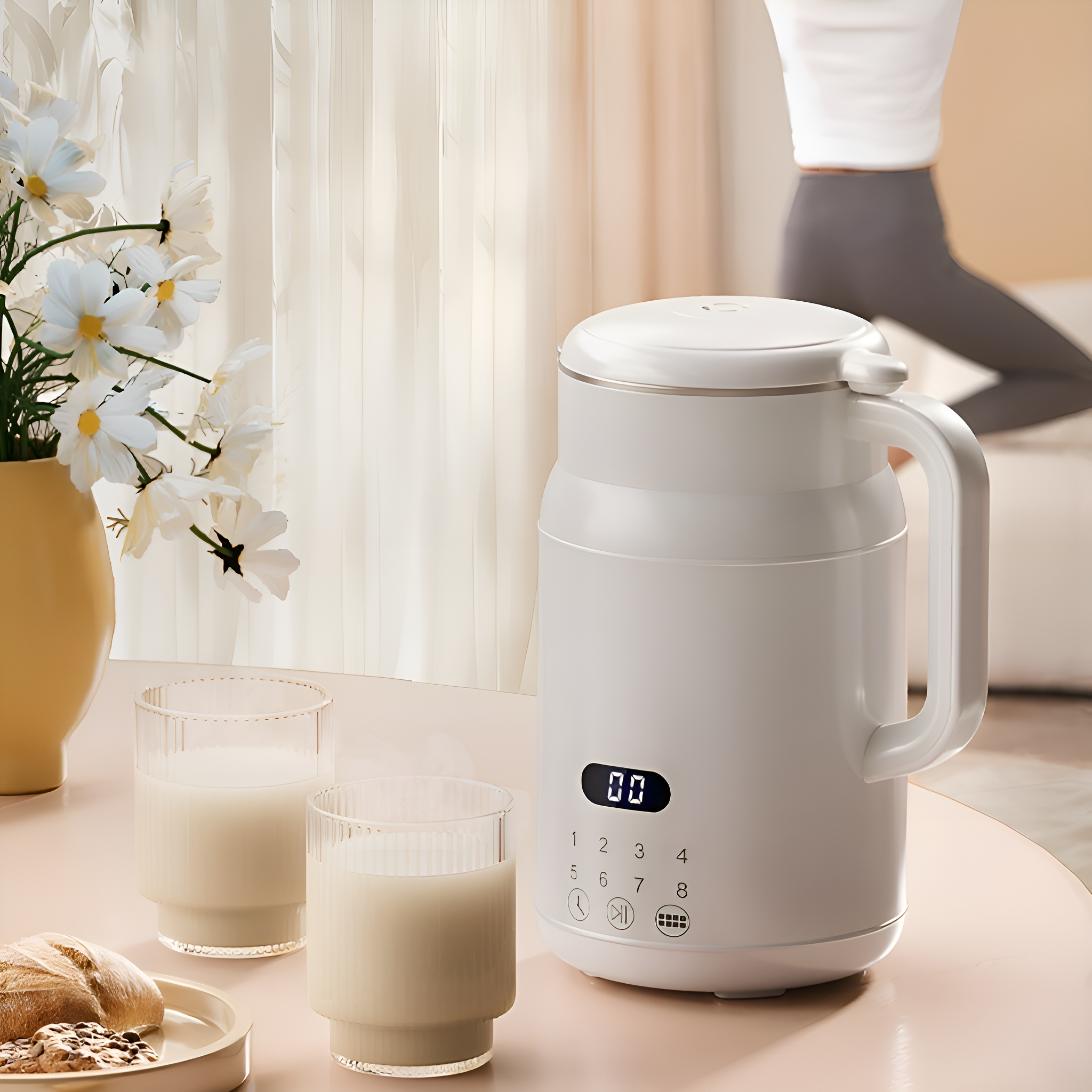 Weißer Sojamilchbereiter auf einem rosa Tisch. Zwei Gläser Milch und Blumen sind in der Nähe.