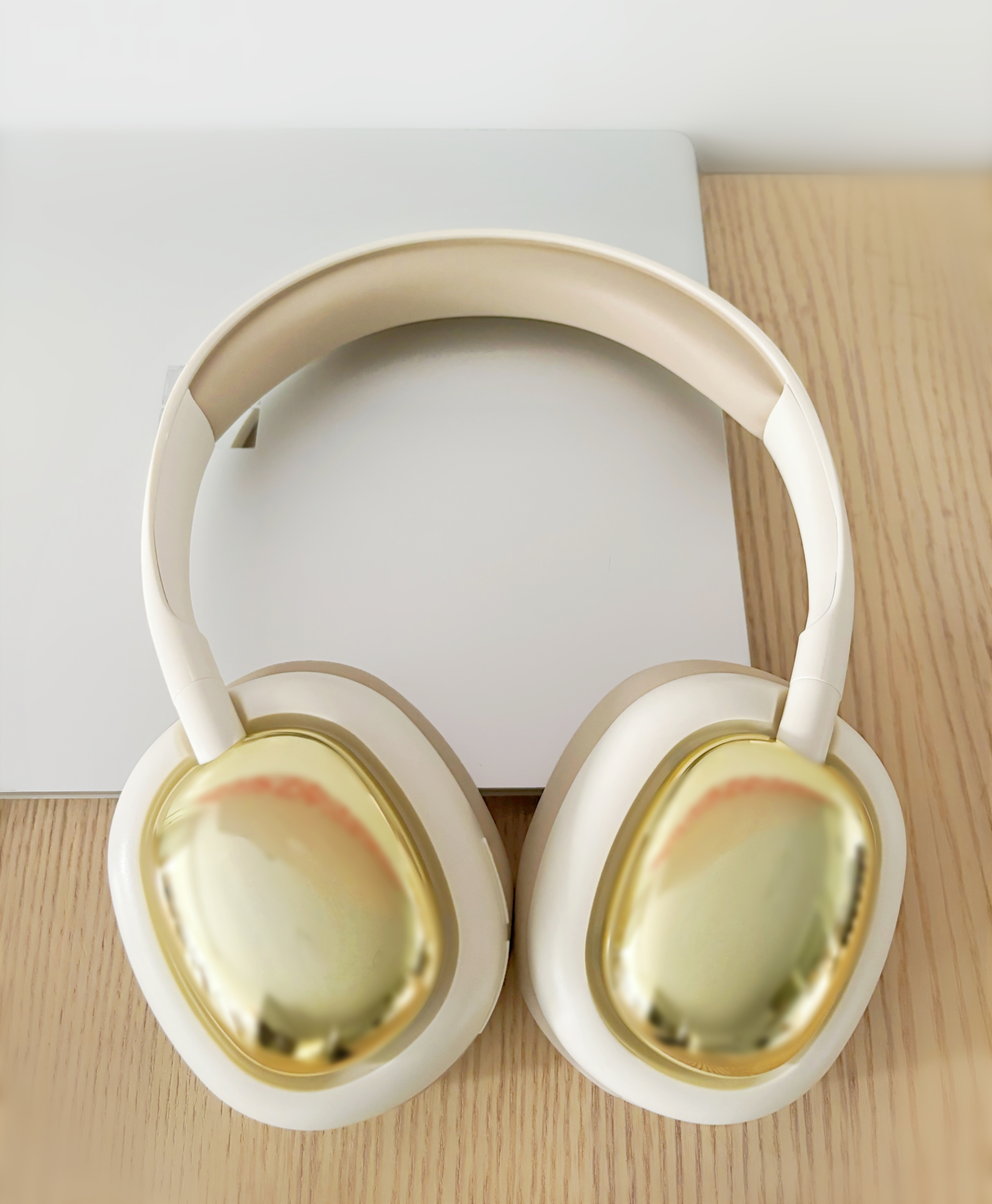 Auriculares blancos y dorados sobre superficie de madera. Portátil al fondo.