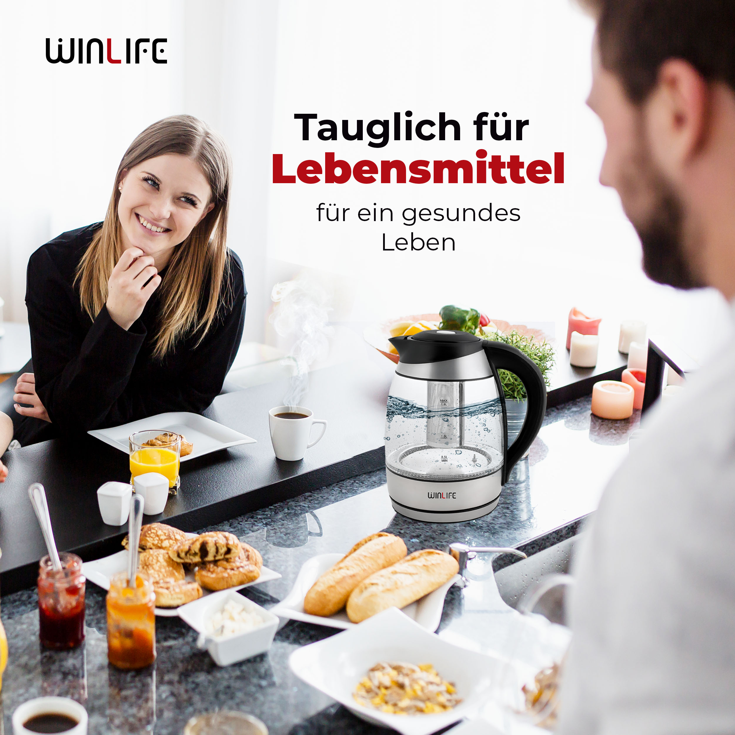 Frau lächelt neben einem Tisch mit Essen und einem Teekessel. Ein Mann schaut auf den Kessel.