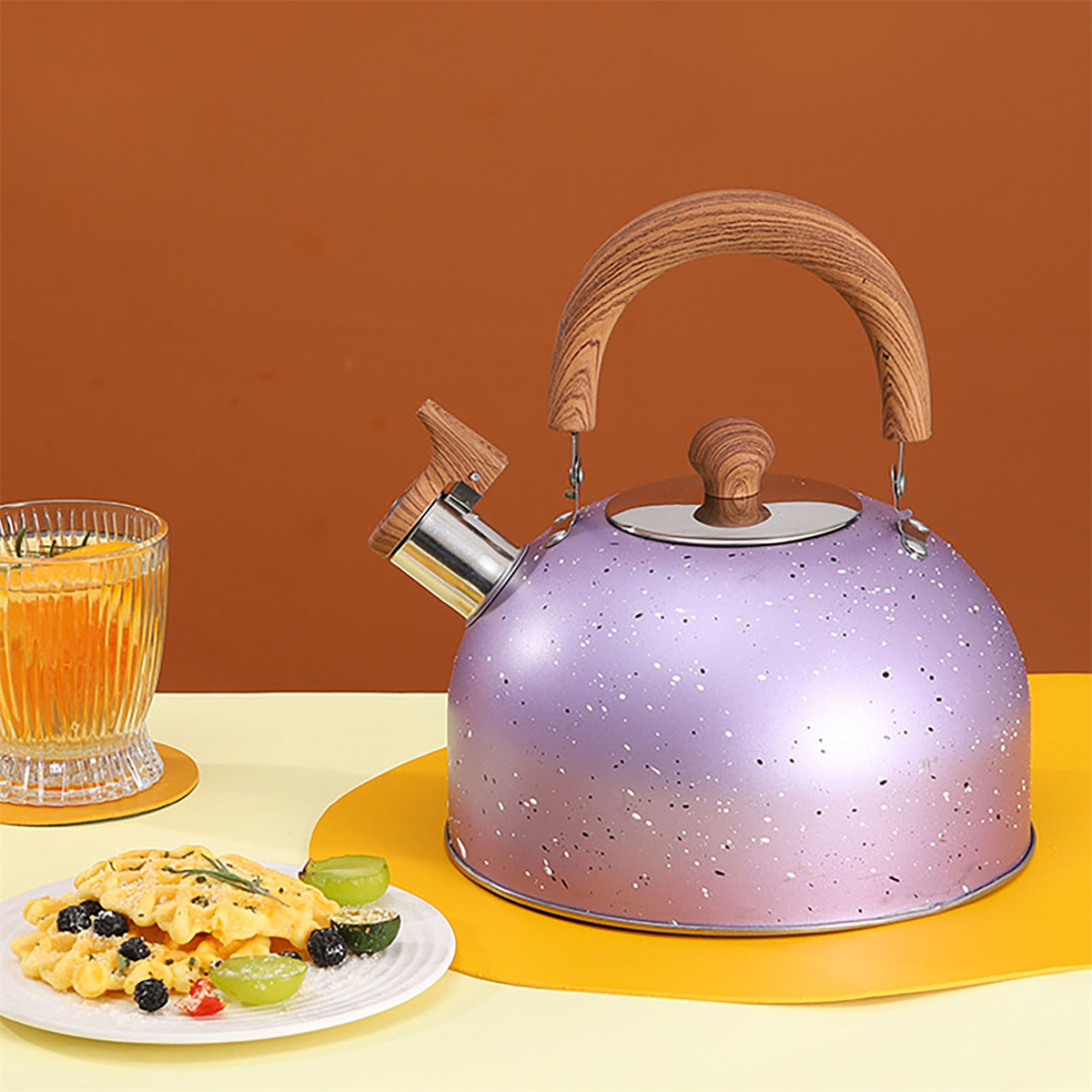 Ein lila gesprenkelter Wasserkocher mit Holzgriff, ein Glas mit orangefarbener Flüssigkeit und ein Teller mit Waffeln und Trauben.